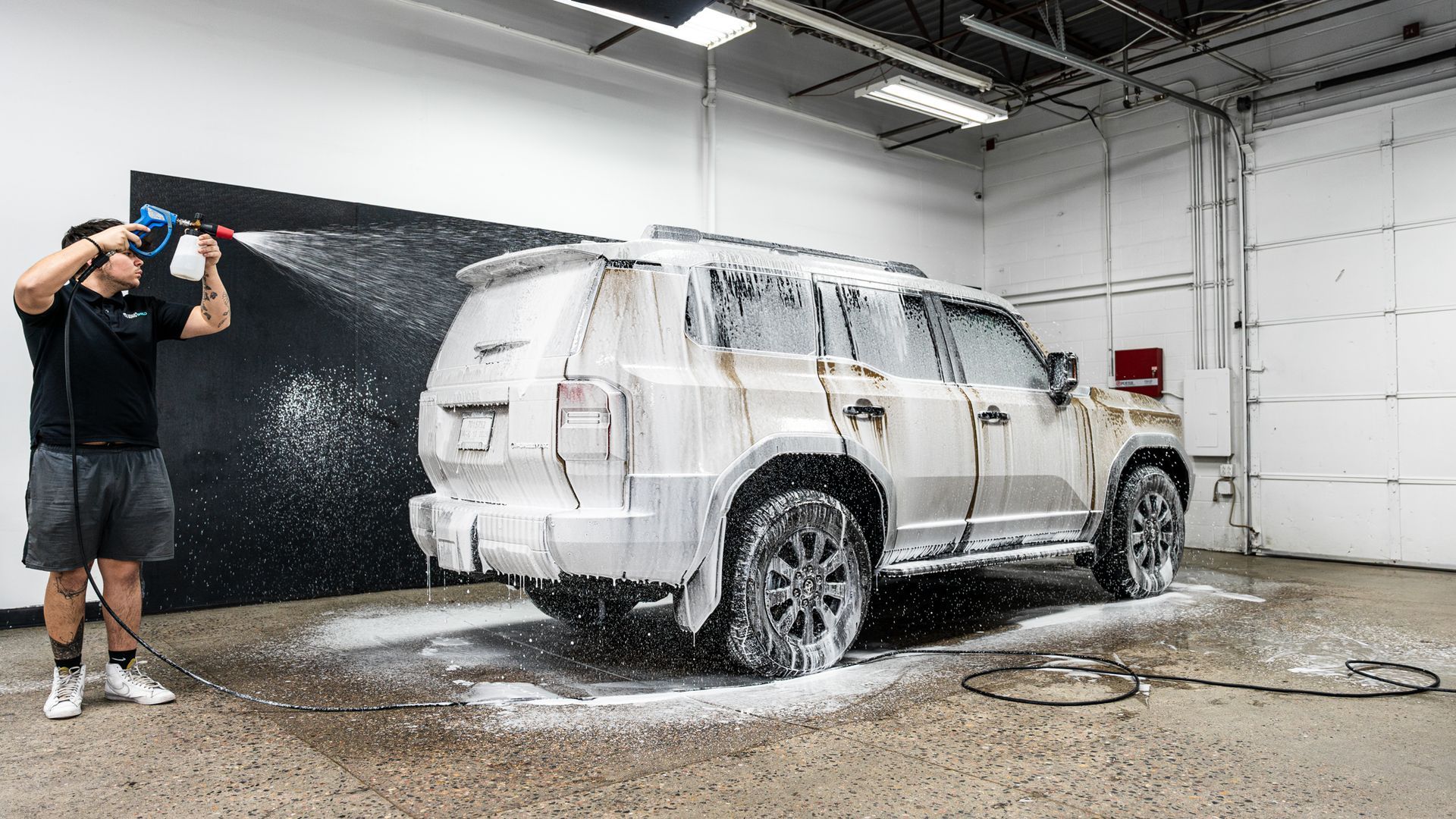 Man spraying a white, soapy foam on a dirty SUV in a car wash bay.