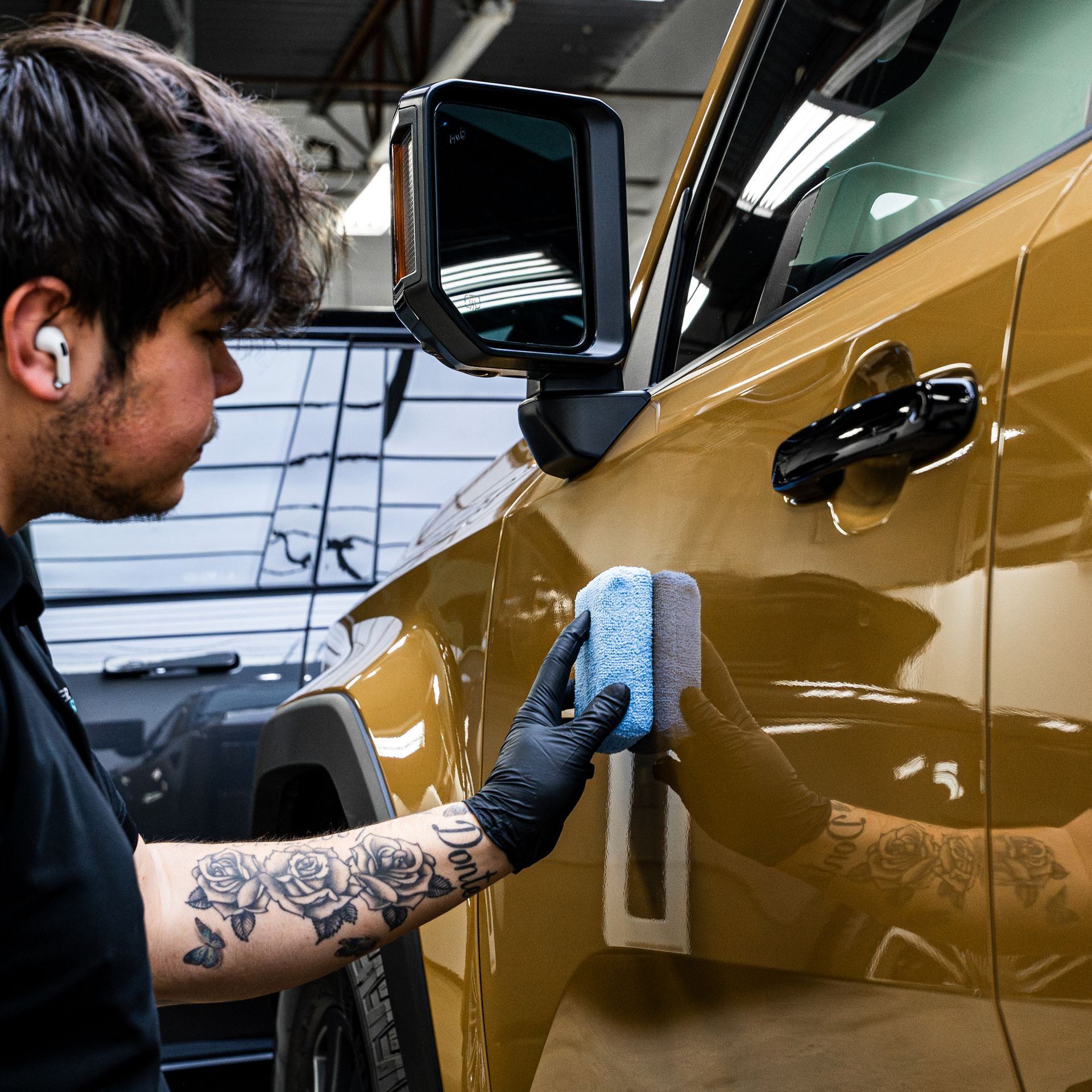 Man applies detailing product to a gold car, wearing black gloves, in a shop.