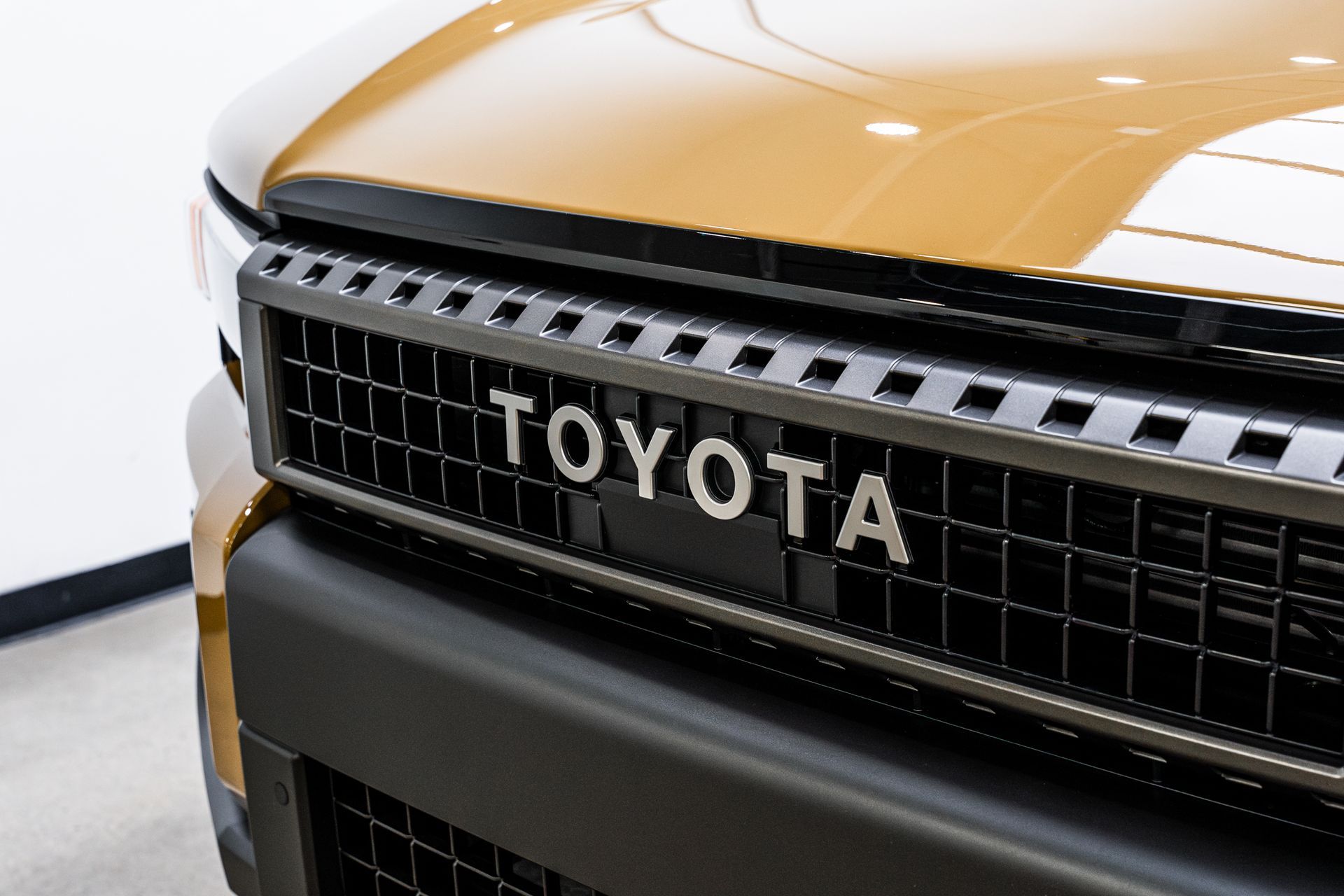 Close-up of a Toyota vehicle's grill. The Toyota logo is centered on a black and silver grill with a tan car body.