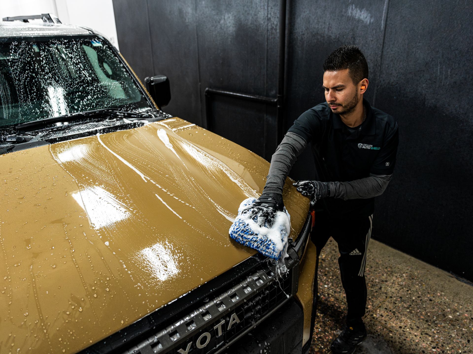 Man washing a tan SUV with soapy sponge in an indoor garage setting.