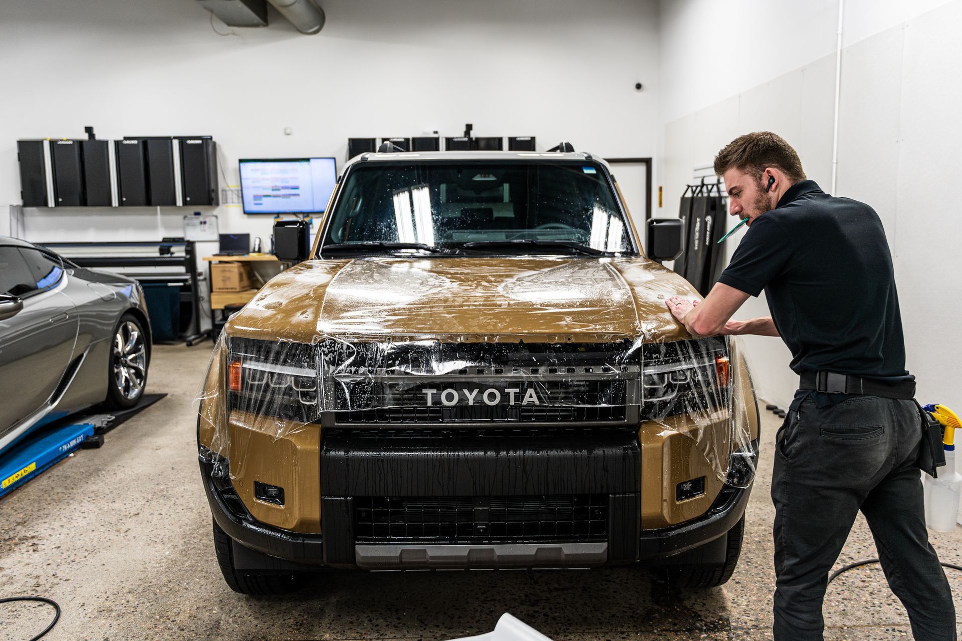 Man applies protective film to a golden Toyota SUV's hood inside a shop.
