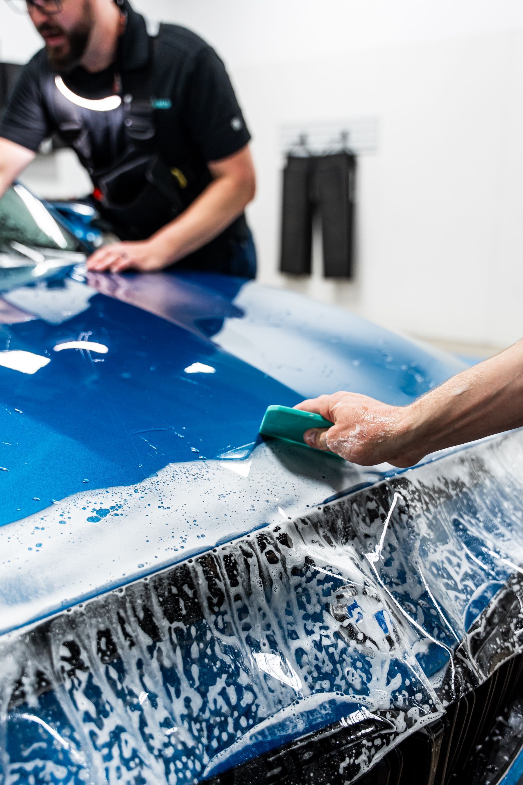 Person using a squeegee to apply film to a blue car hood, soapy water visible, another person nearby.