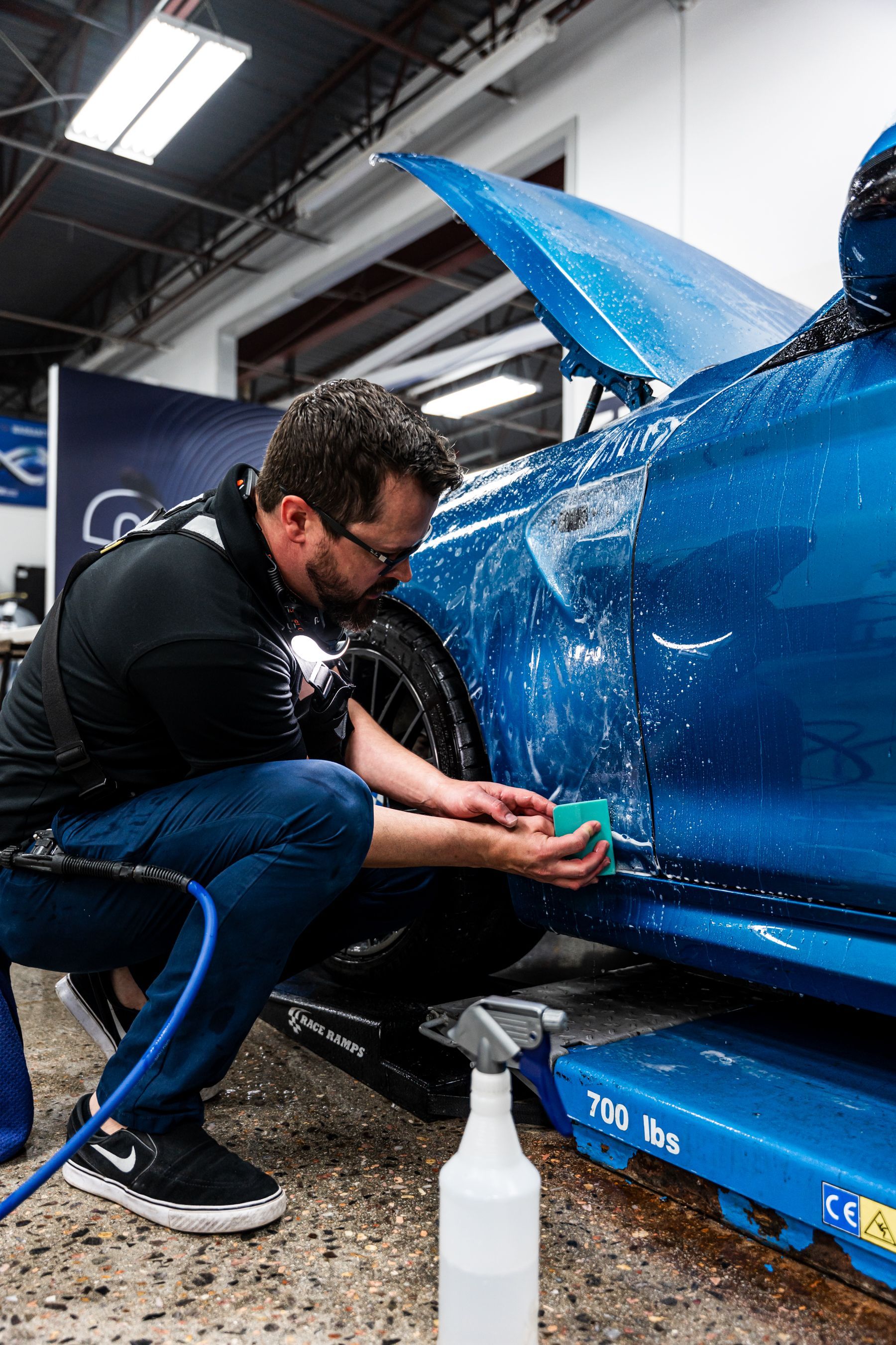 Man washes blue car with a sponge, water spraying. Shop setting, squatting, focused expression.