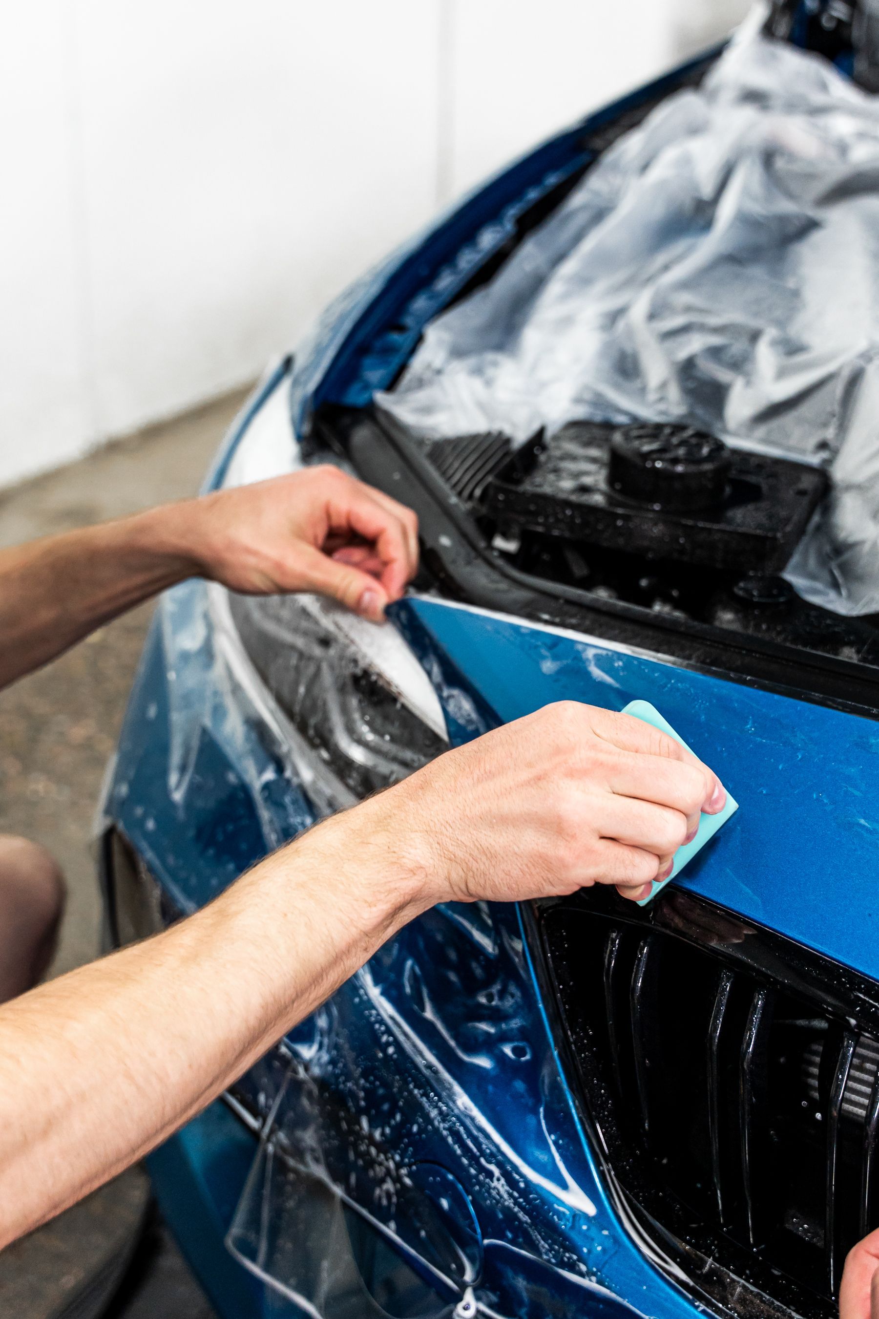 Person applying protective film to a blue car's hood with a sponge in a garage.