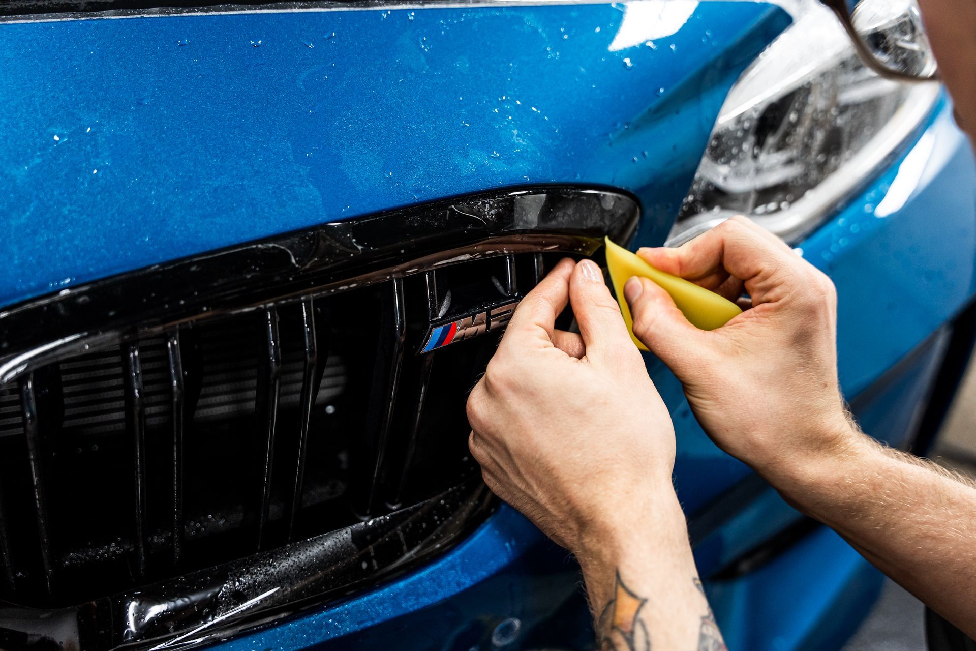 Person applying blue protective film to a black car grill with a yellow squeegee.