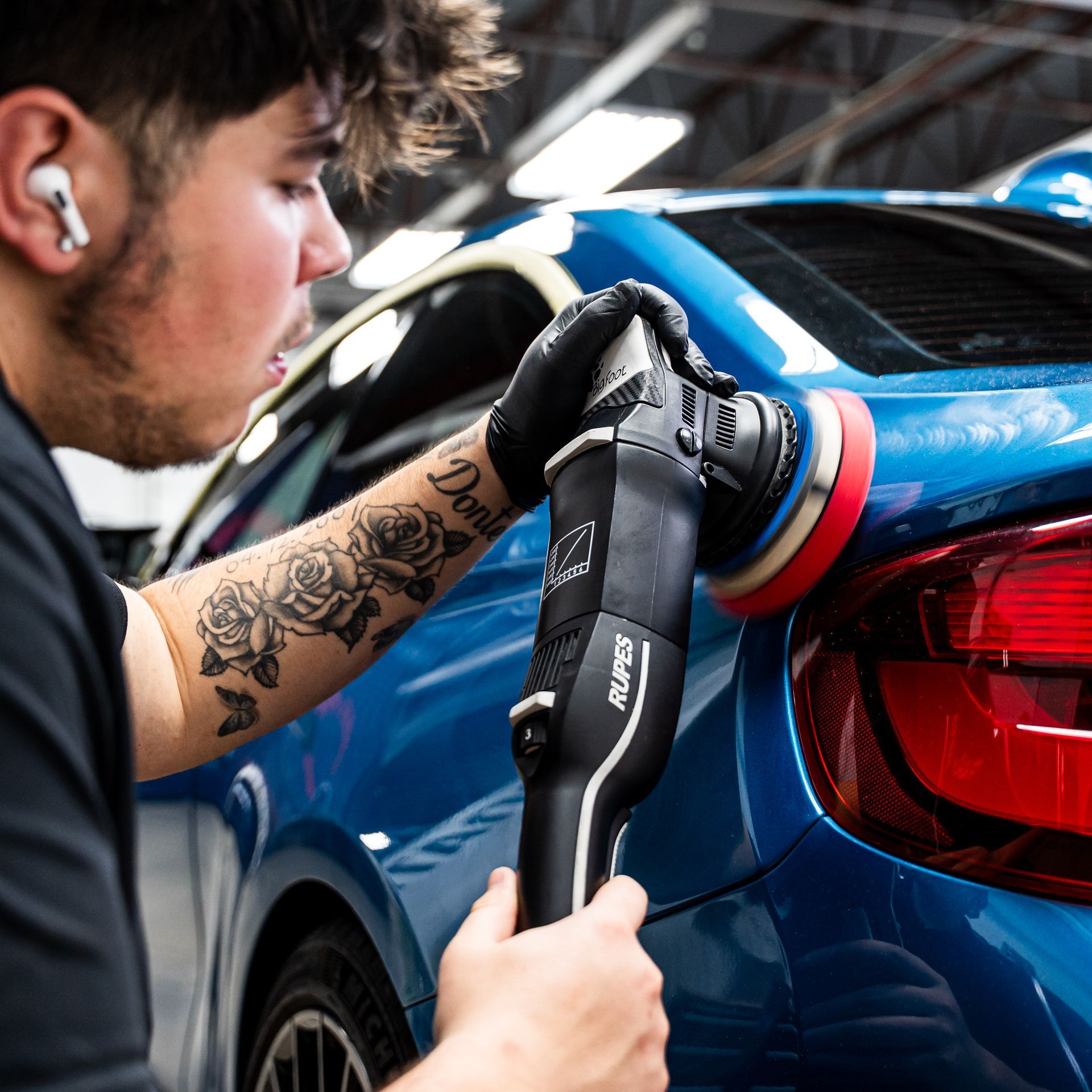 Man polishing a blue car with a machine buffer, wearing gloves, in a garage.