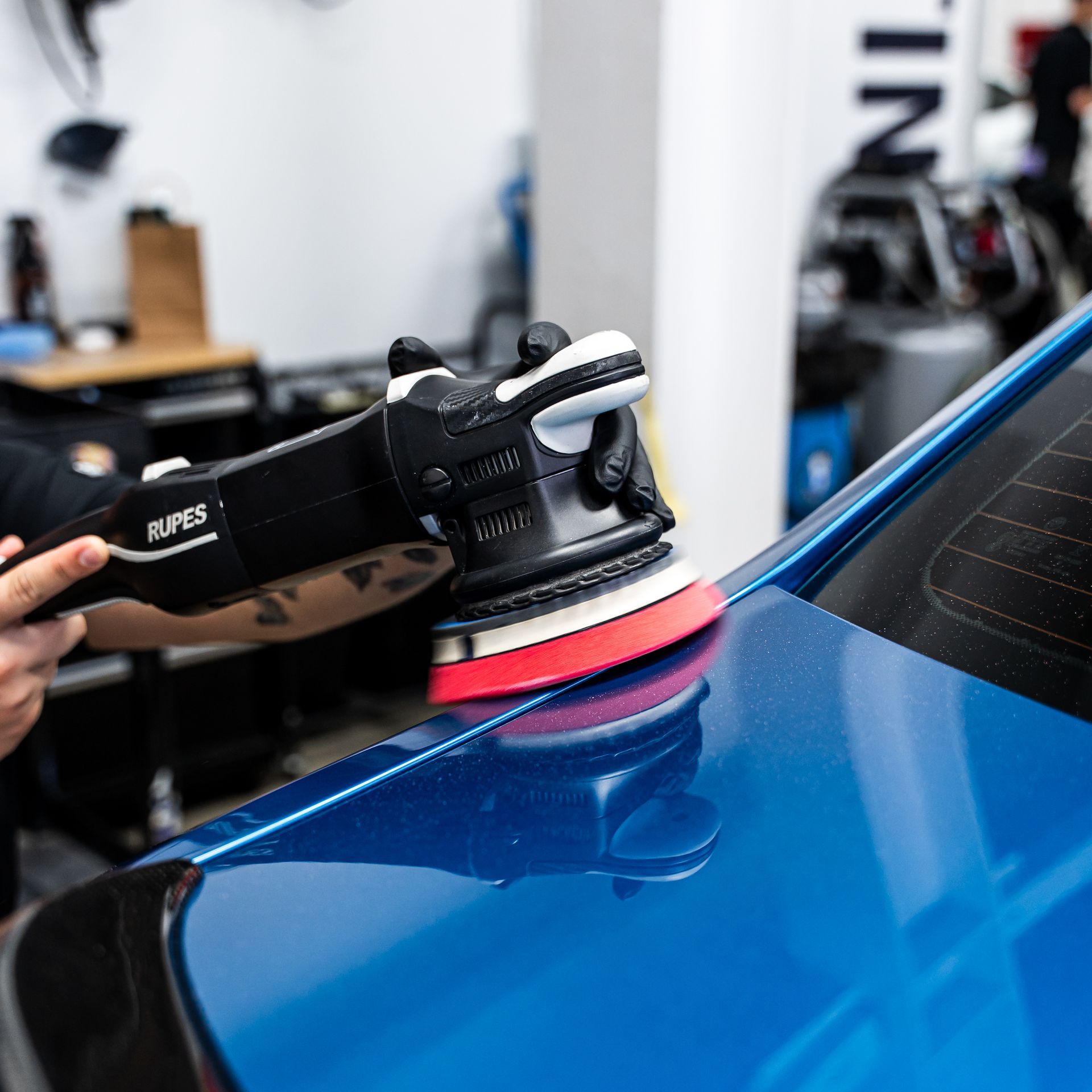 Person polishing blue car with a black and white polisher in a shop.