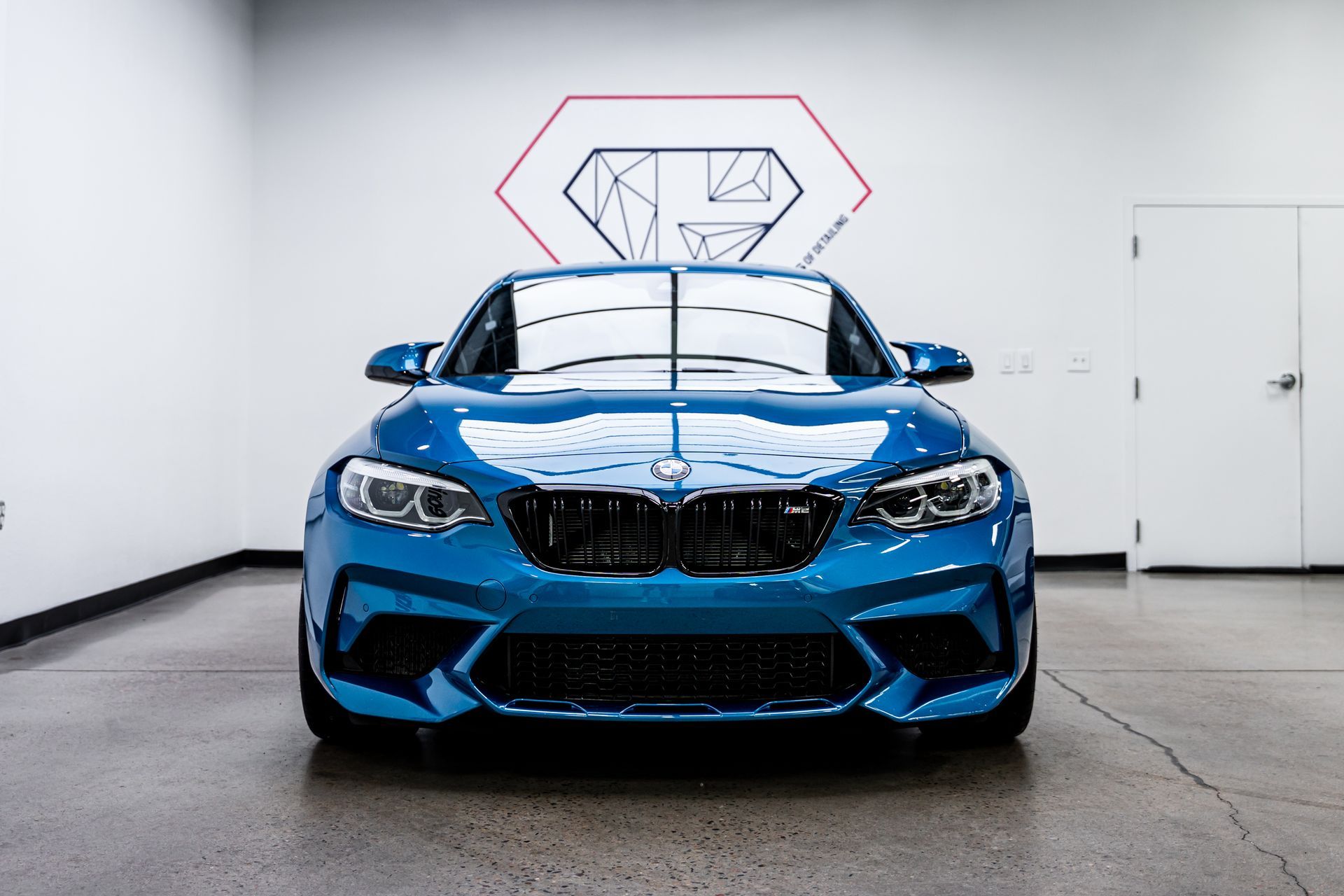 Blue BMW M2 in a white garage, facing forward.