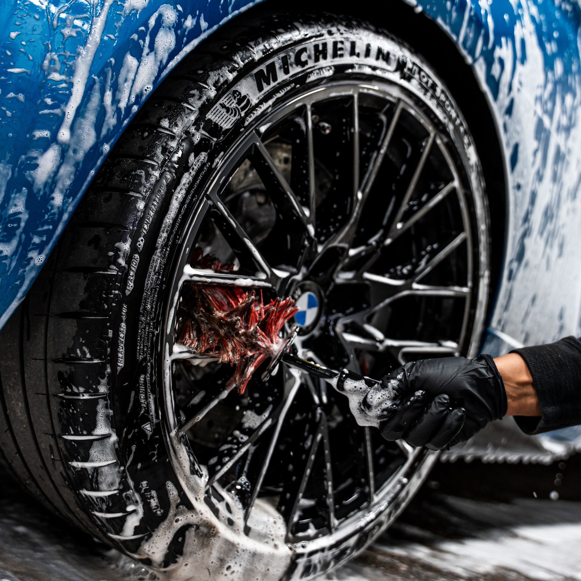 Blue car wheel being washed with soapy water; black gloved hand scrubbing.