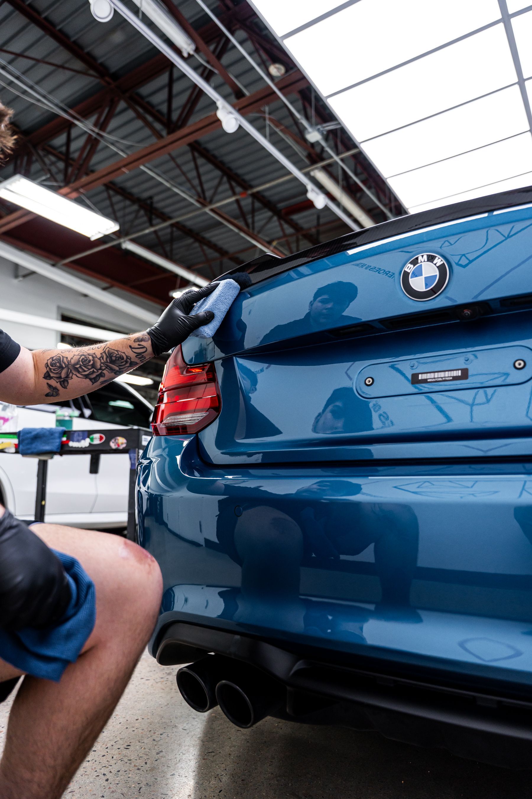 Person cleaning a blue BMW's rear with a microfiber cloth, inside a well-lit shop.