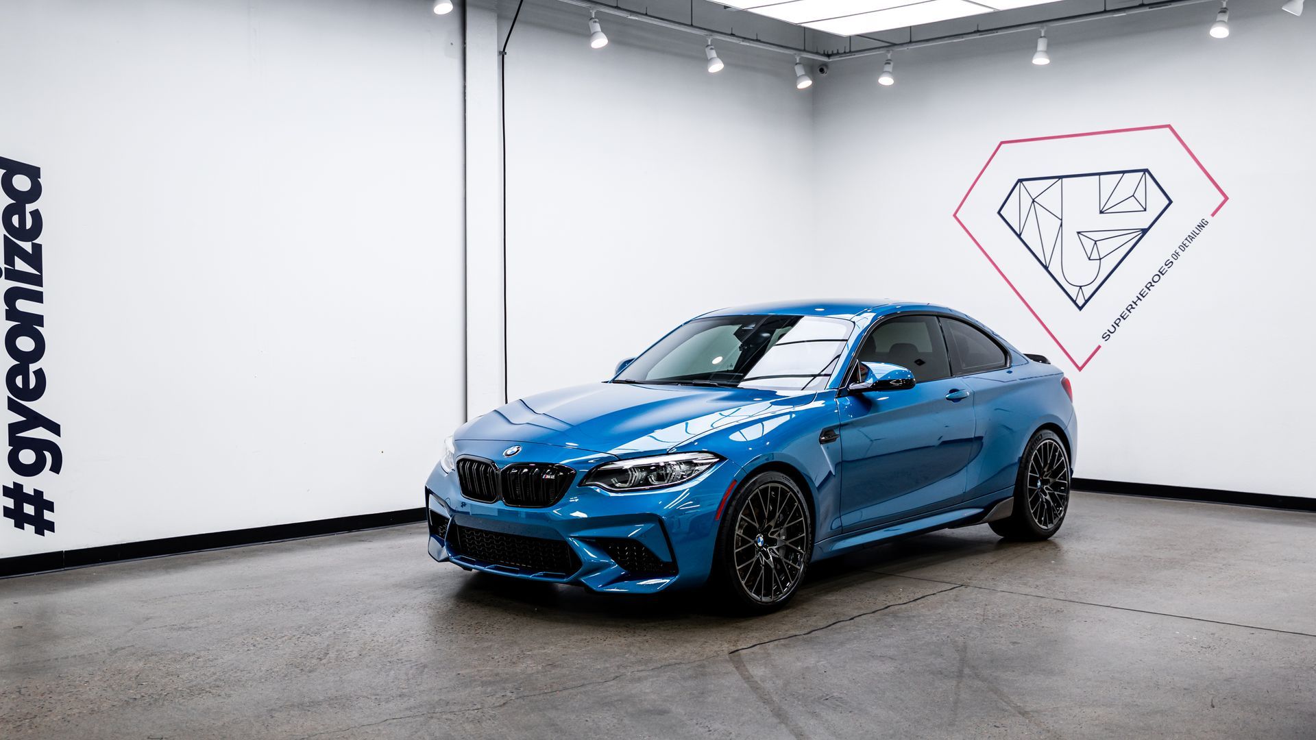 Blue BMW M2 coupe in a well-lit showroom. Diamond logo on wall, #gyeonized on the left.