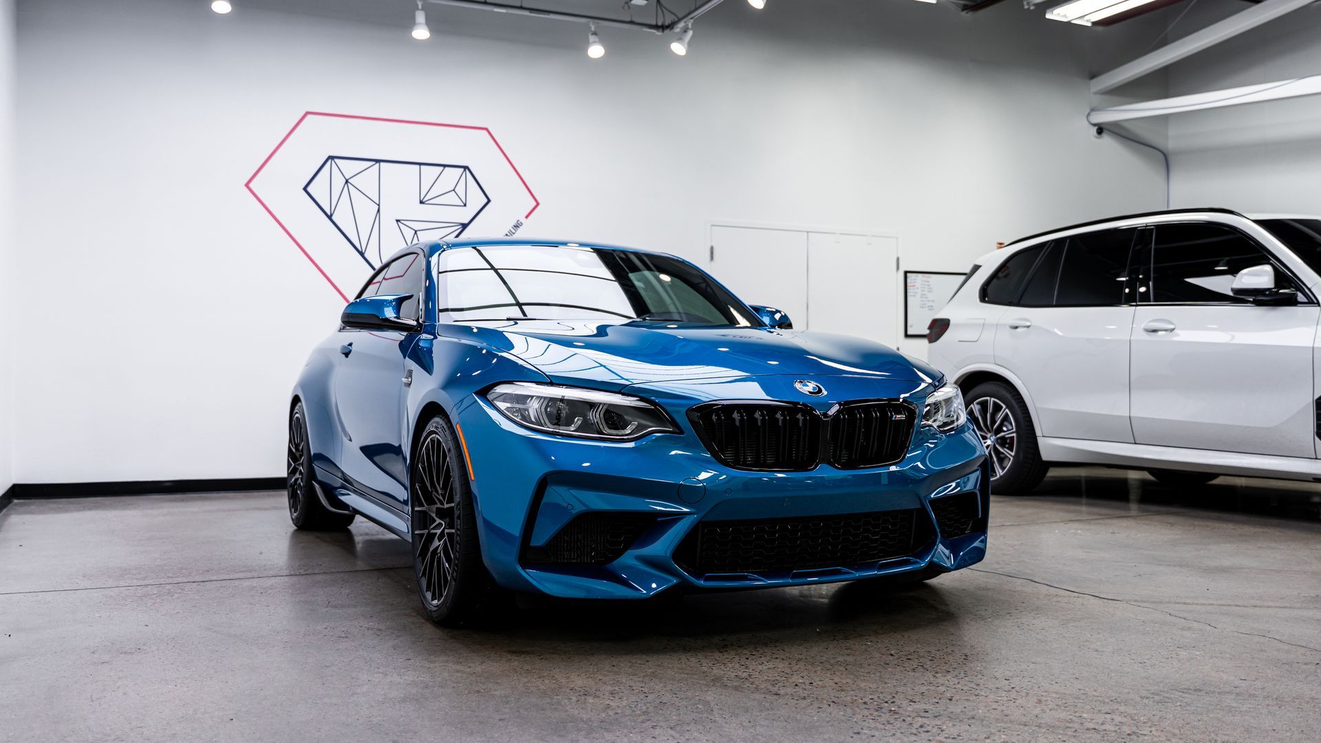 Blue BMW M2 coupe parked inside a showroom, with a white SUV in the background.