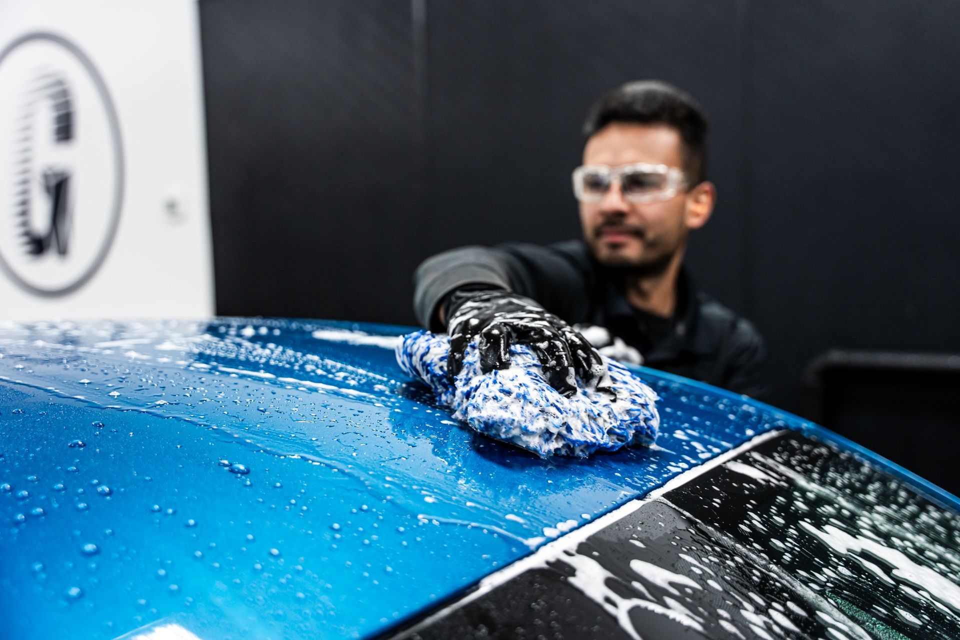 Man washing a blue car, wearing safety glasses and gloves, indoors with black wall.