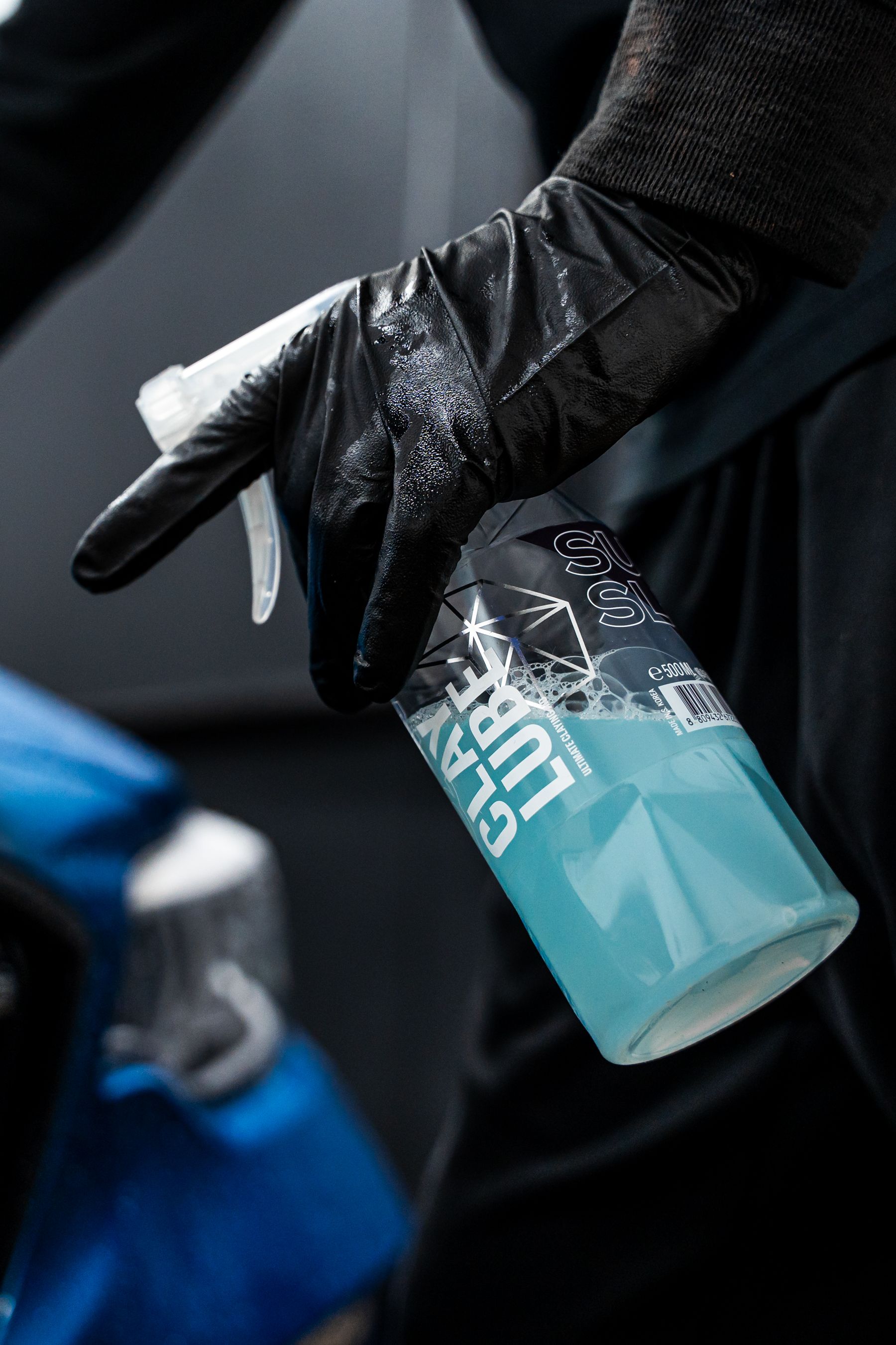 Gloved hand holding a spray bottle of clay lubricant, spraying towards a blue car.