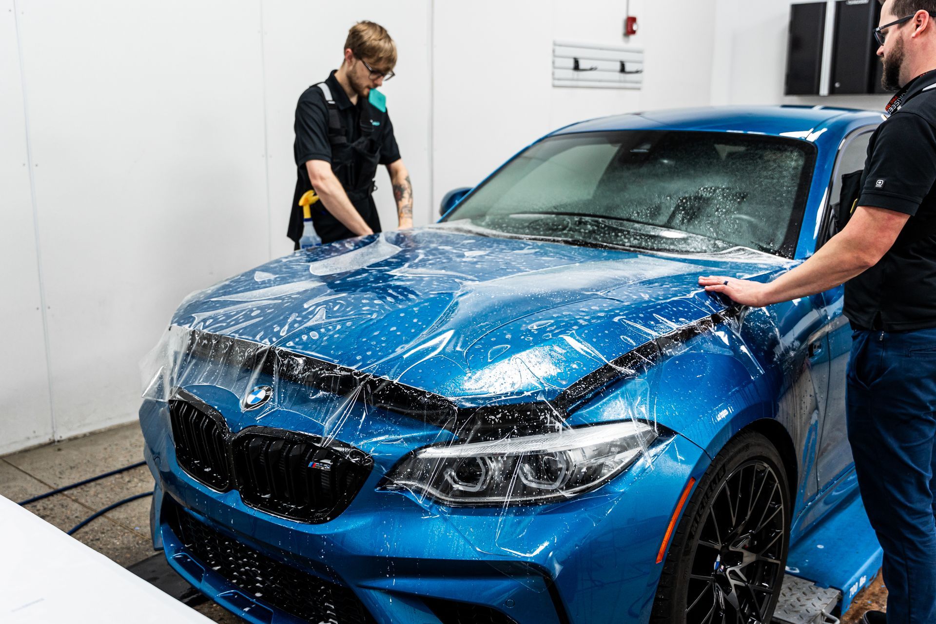 Two people applying protective film to a blue car's hood in a shop.