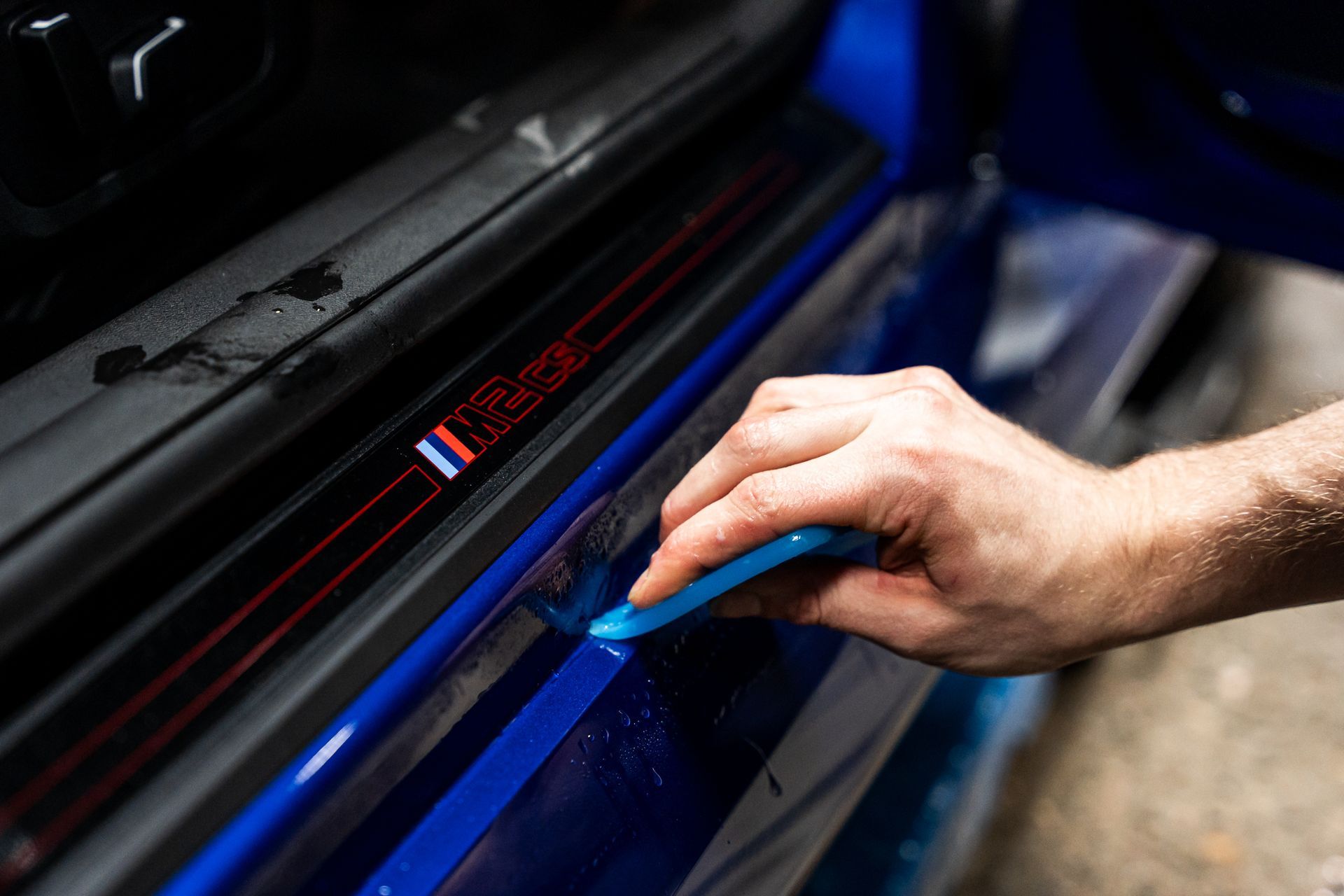 Person using a blue tool to apply a protective film to a car door sill. Red trim and