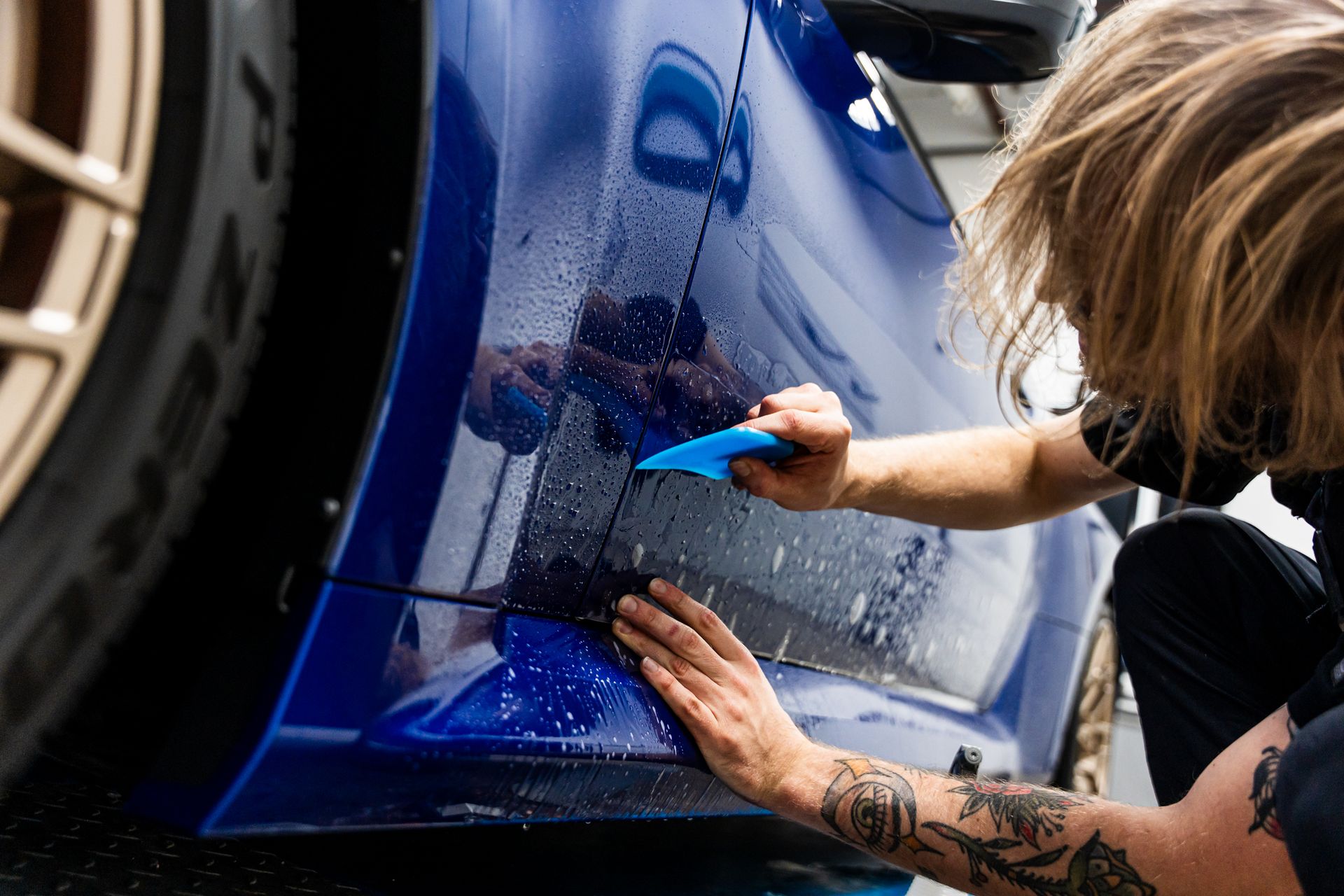 Person applying protective film to a blue car bumper with a squeegee.