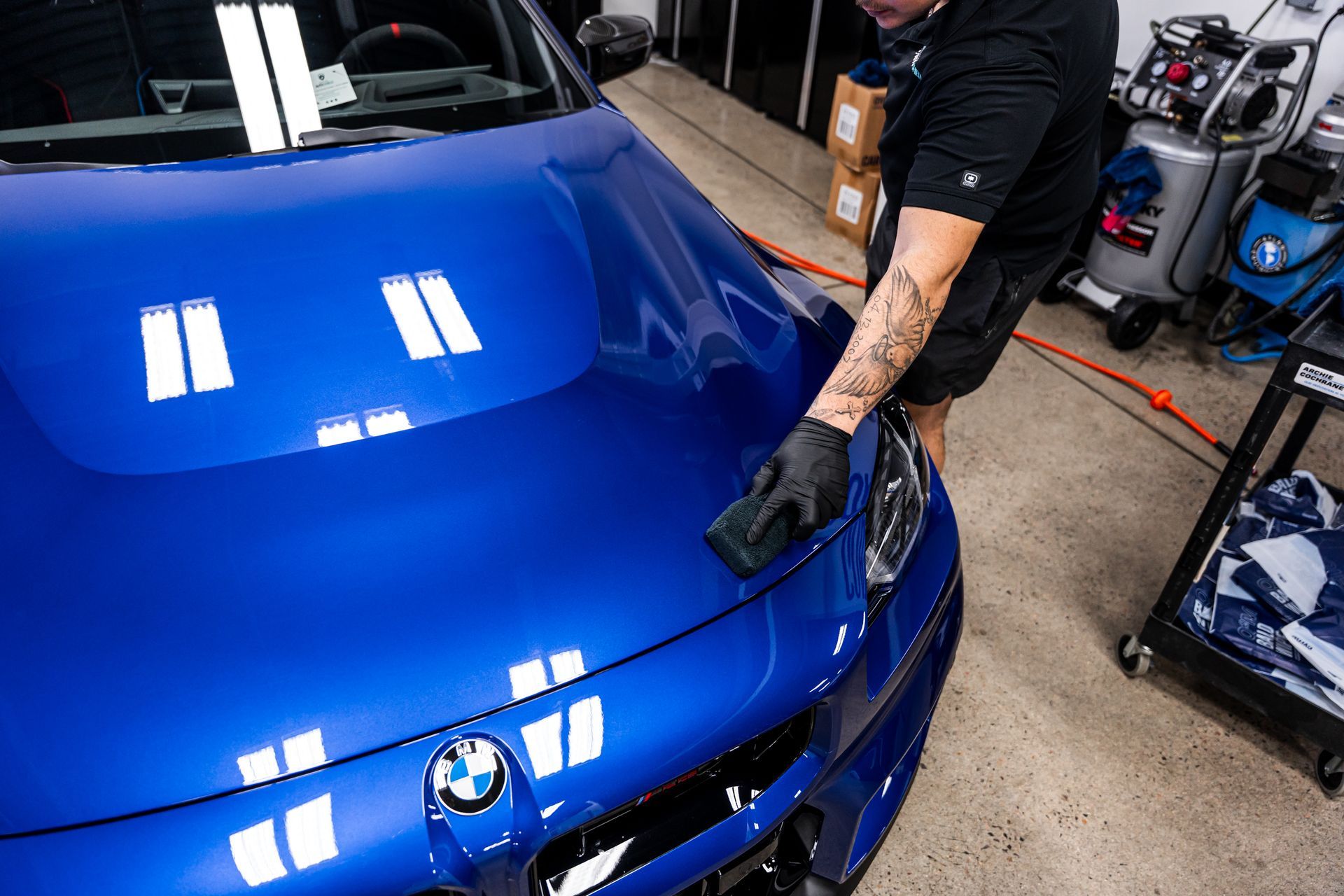 A person wearing black gloves is detailing a blue BMW car hood in a garage.