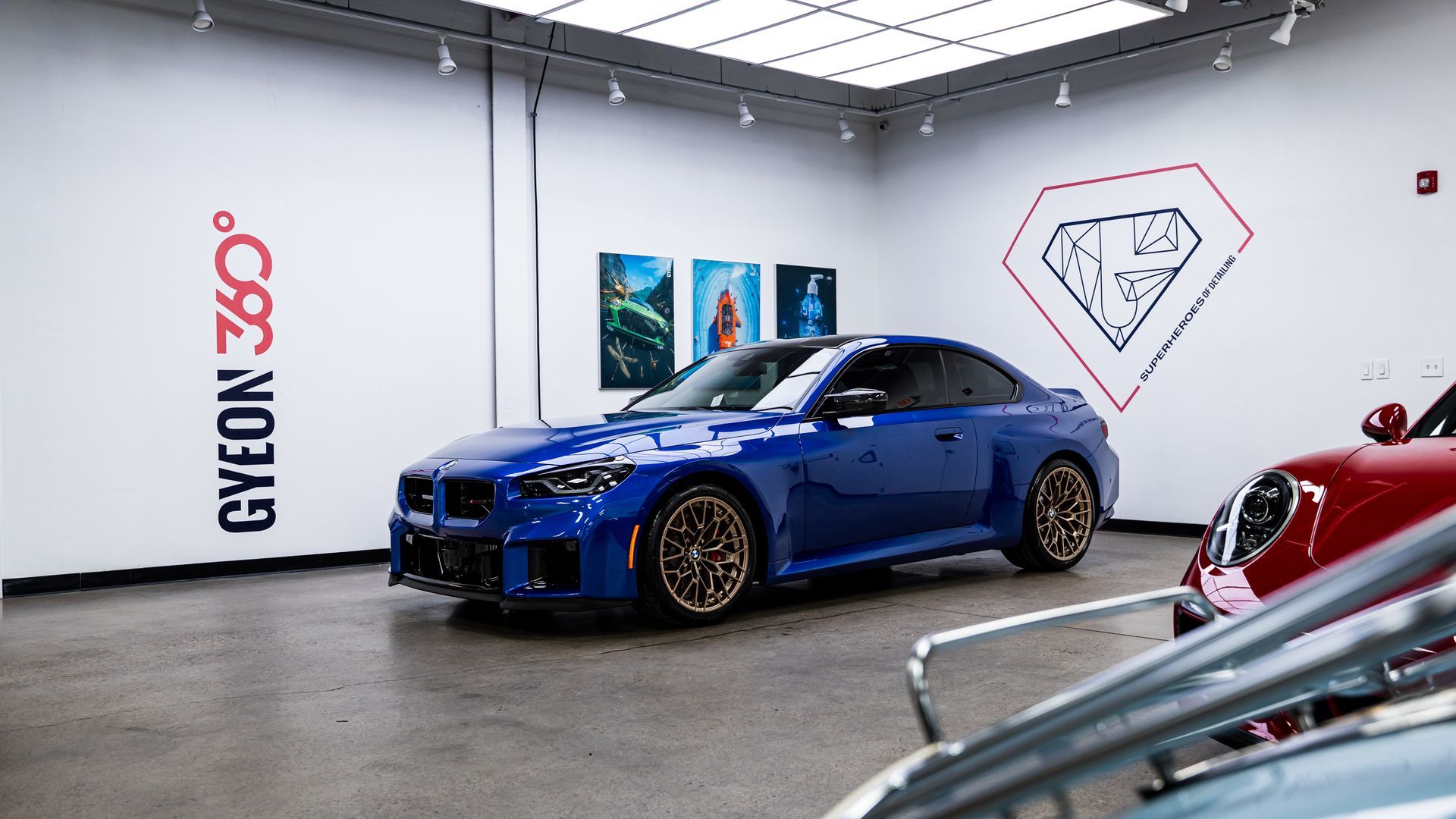 Blue sports car in a clean, white garage. Another red car is partially visible. Gyeon 360 logo on wall.