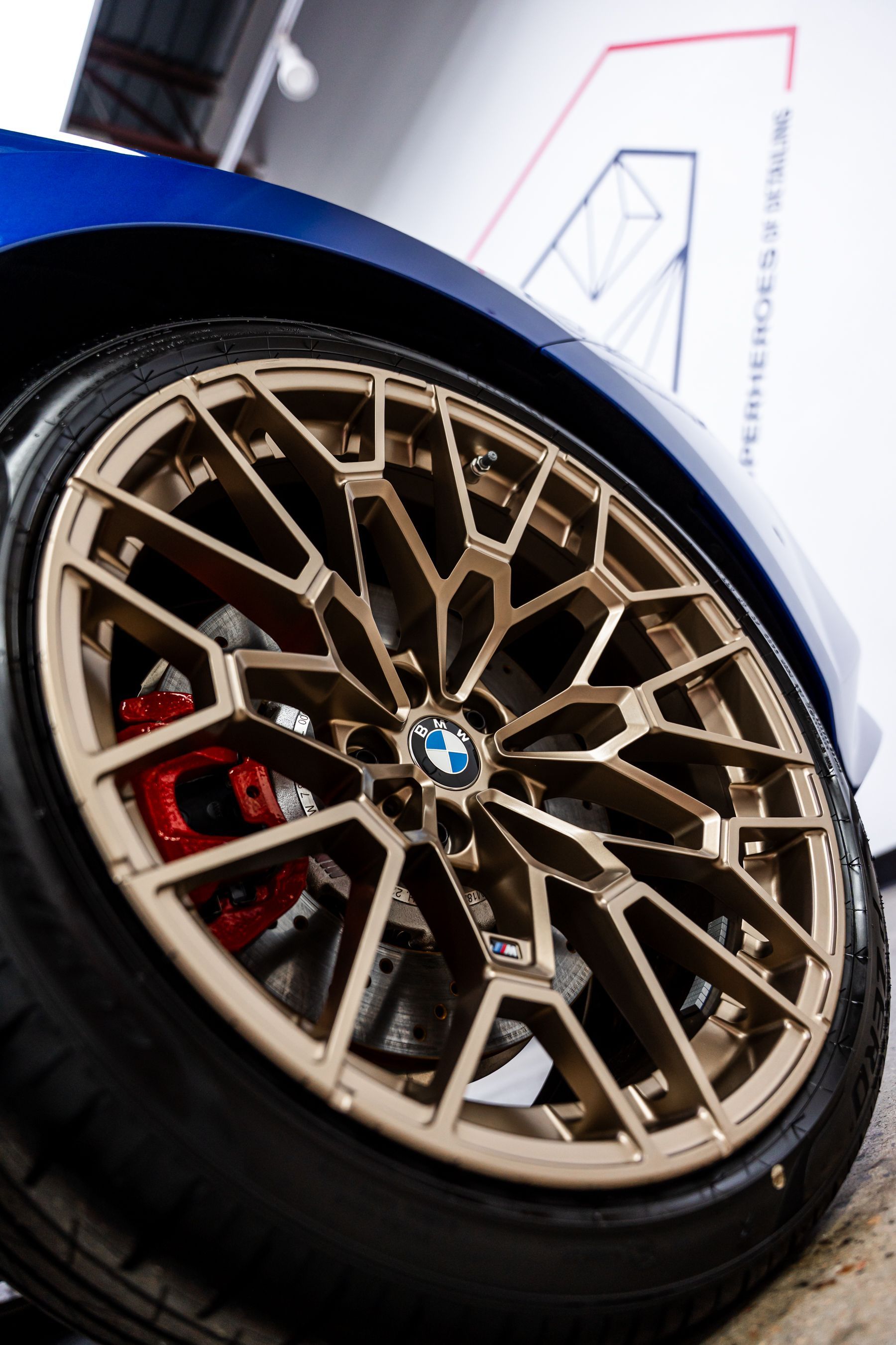 Gold BMW wheel with red brake calipers, blue car body, indoors.