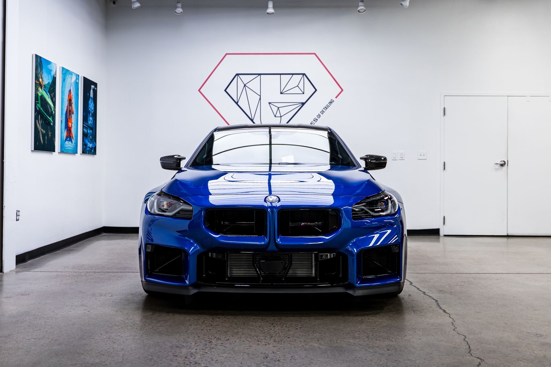 Blue sports car parked in a showroom, under a geometric logo, with art on the wall.