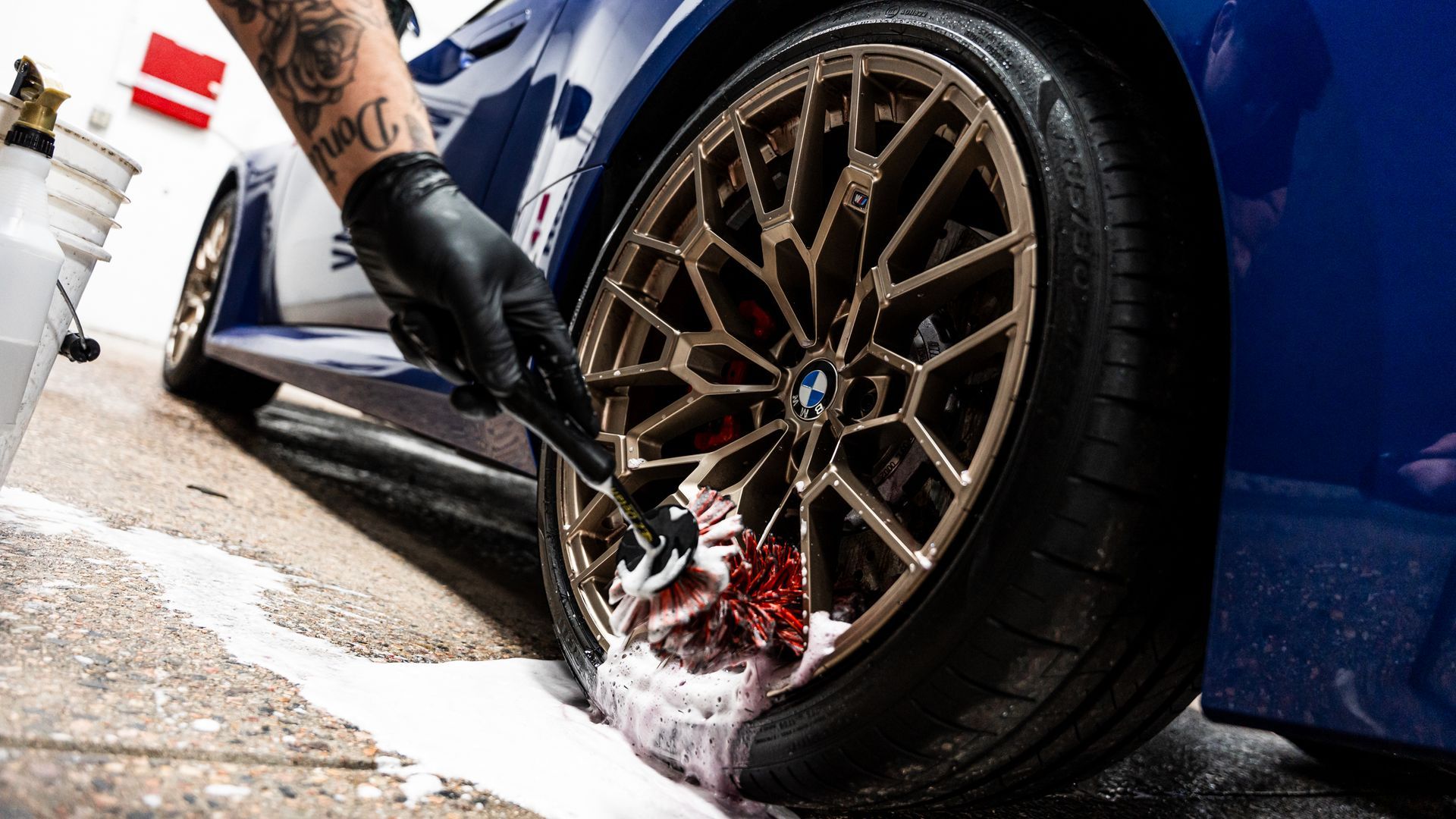 Person washing the bronze wheel of a blue BMW with soapy water, wearing a black glove.