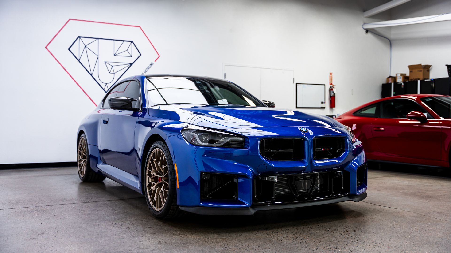 Blue BMW coupe with bronze wheels inside a garage; red car in background, logo on wall.