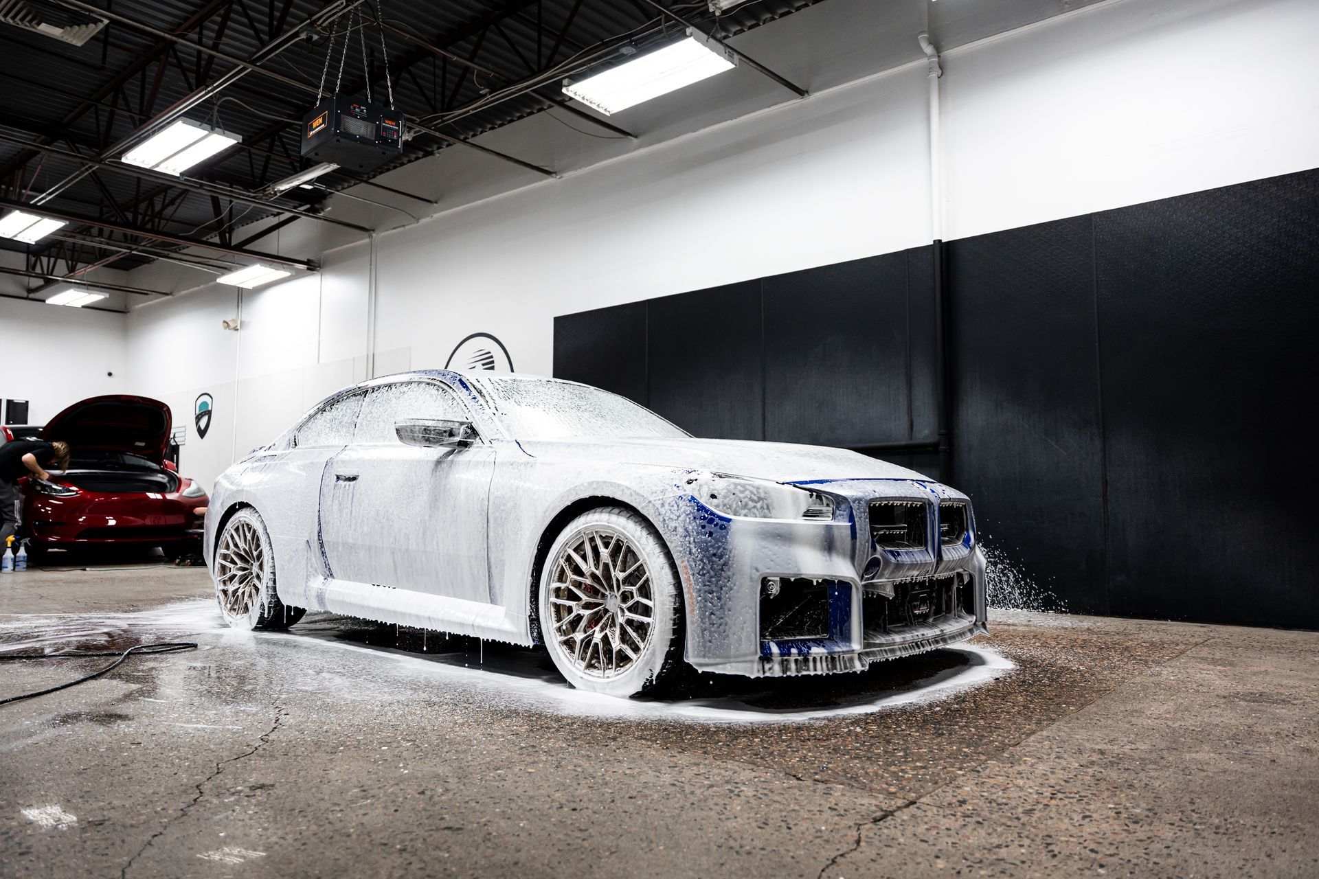 Car covered in white foam at a car wash. A person is visible by a red car in the background.