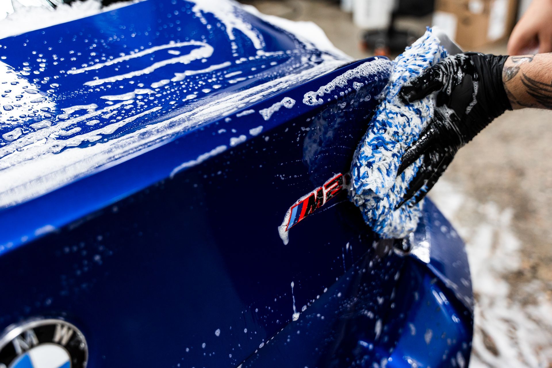 Person washing a blue BMW car with a soapy sponge, close-up on the trunk with the M badge.