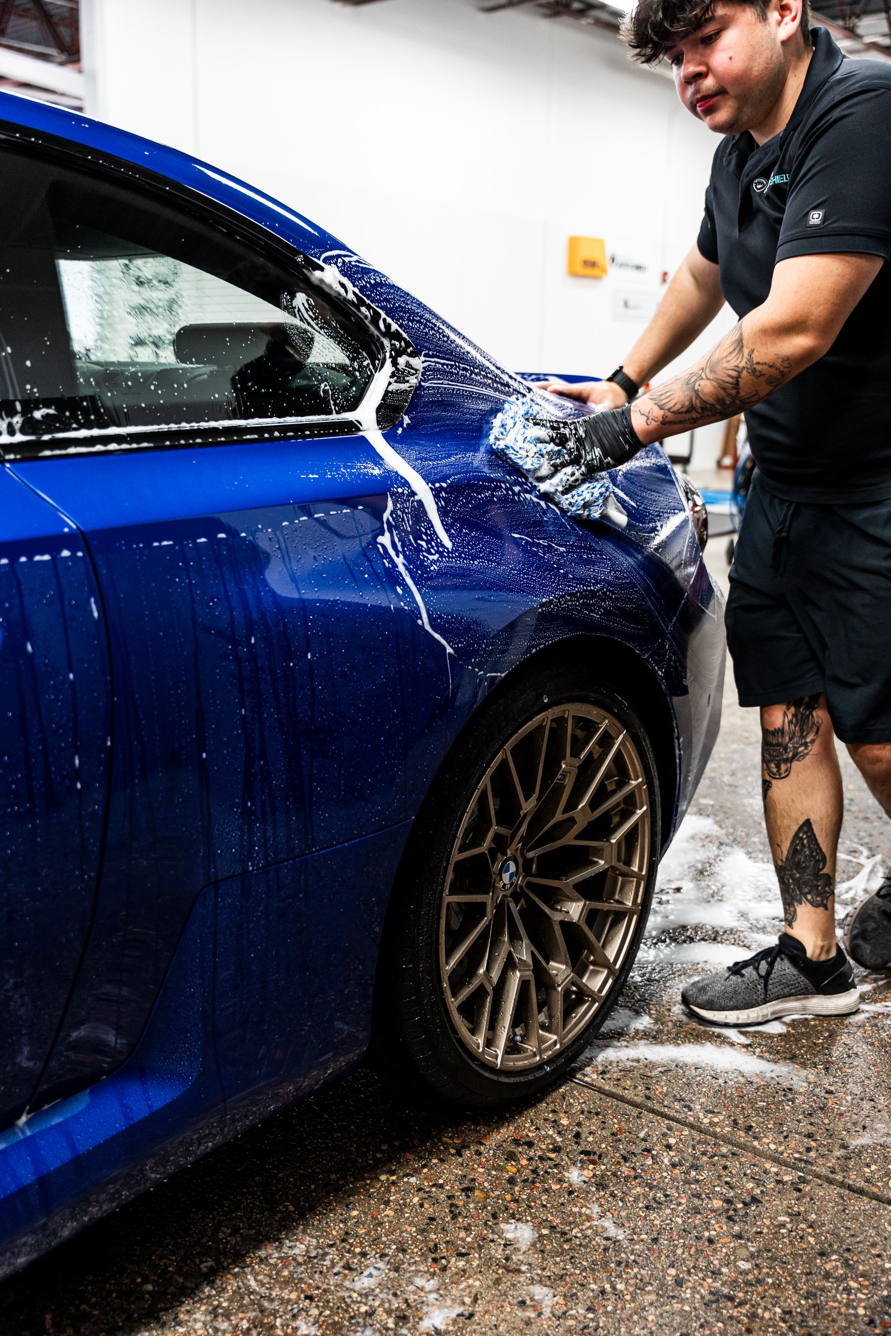 Person washing a blue car with soapy water; gold wheel visible.