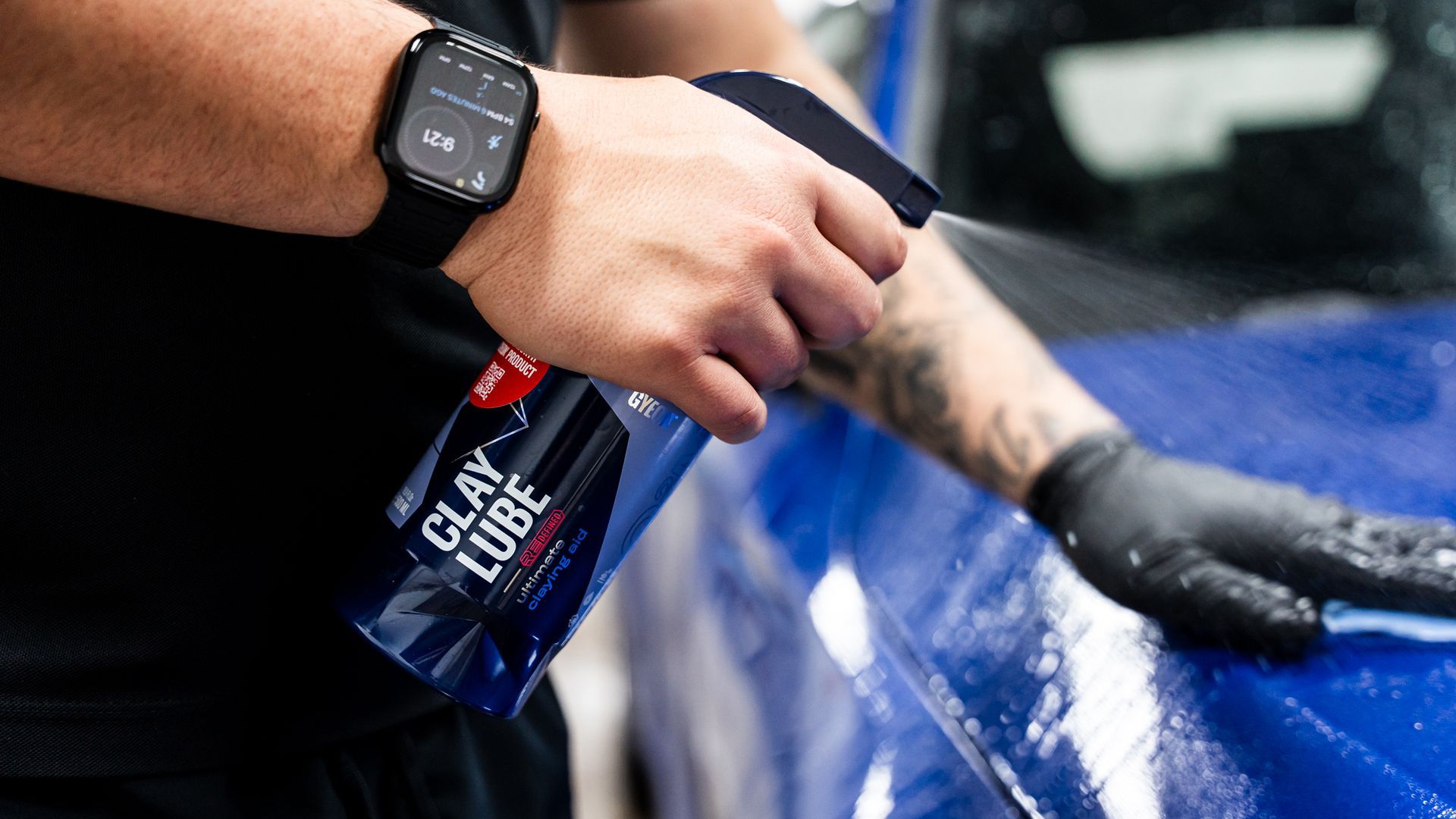 Person spraying blue clay lube on a car's surface, wearing a watch and black gloves.
