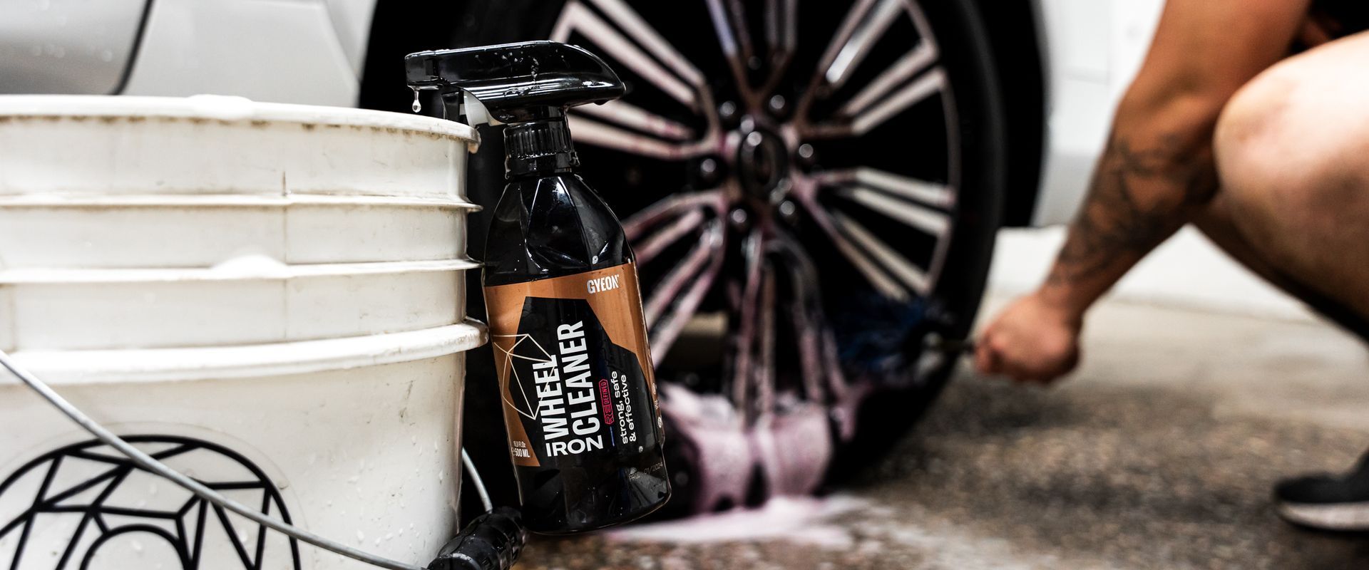 Person cleaning a car wheel with spray and a bucket, wheel cleaner product visible.