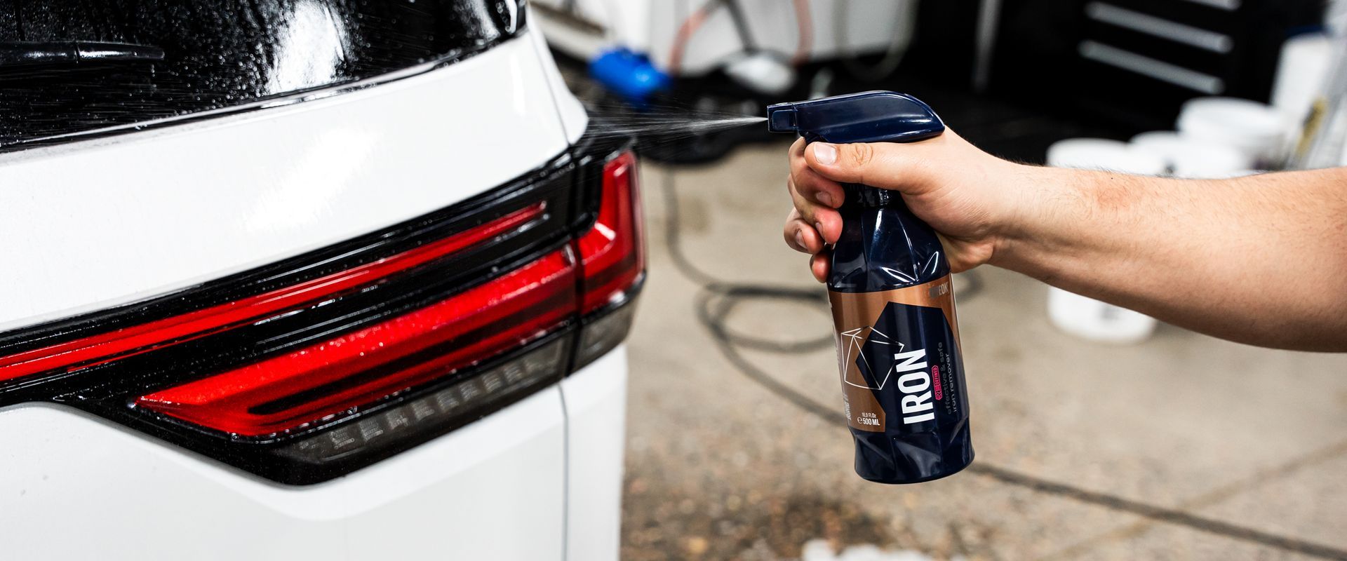 A hand sprays a bottle of car detailing product on a white car's taillight.