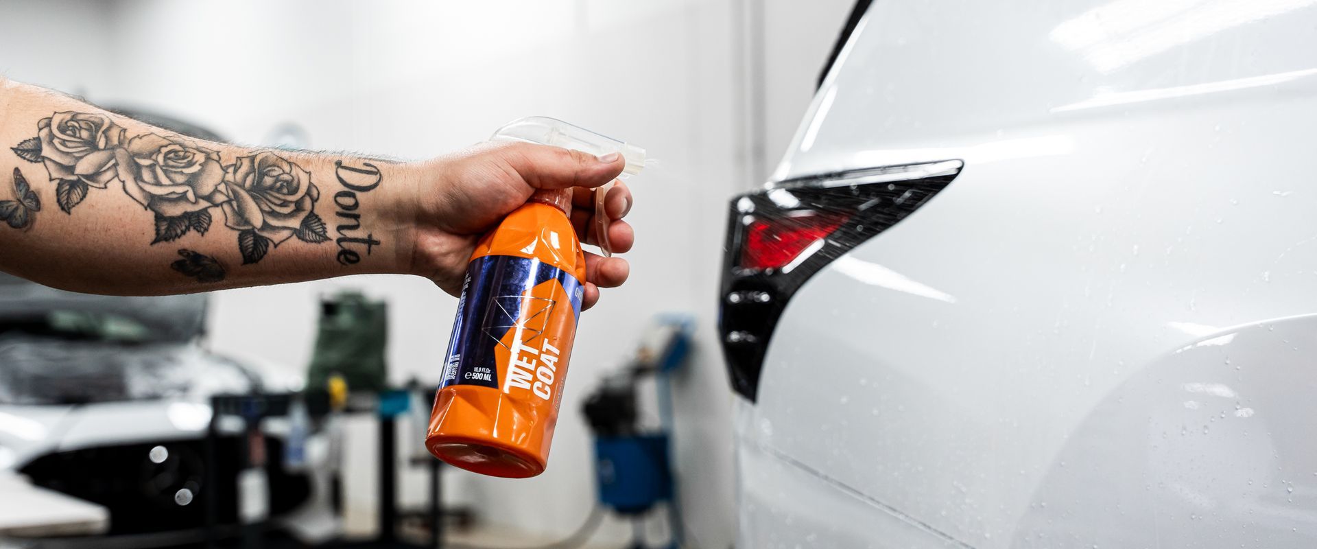 A person with rose tattoos sprays a white car with an orange bottle in a garage.