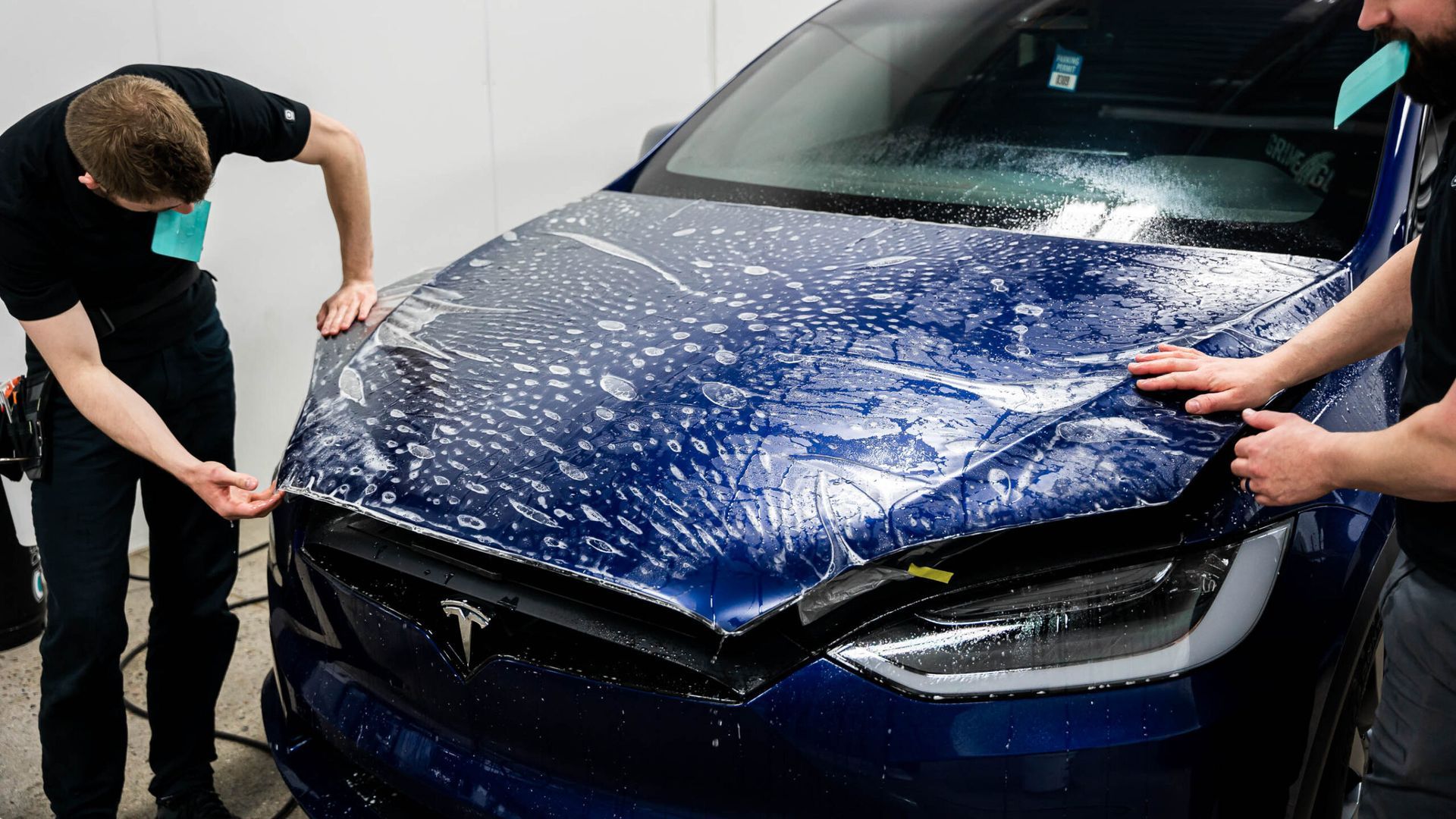 Two men are wrapping a blue tesla model x with a protective film.