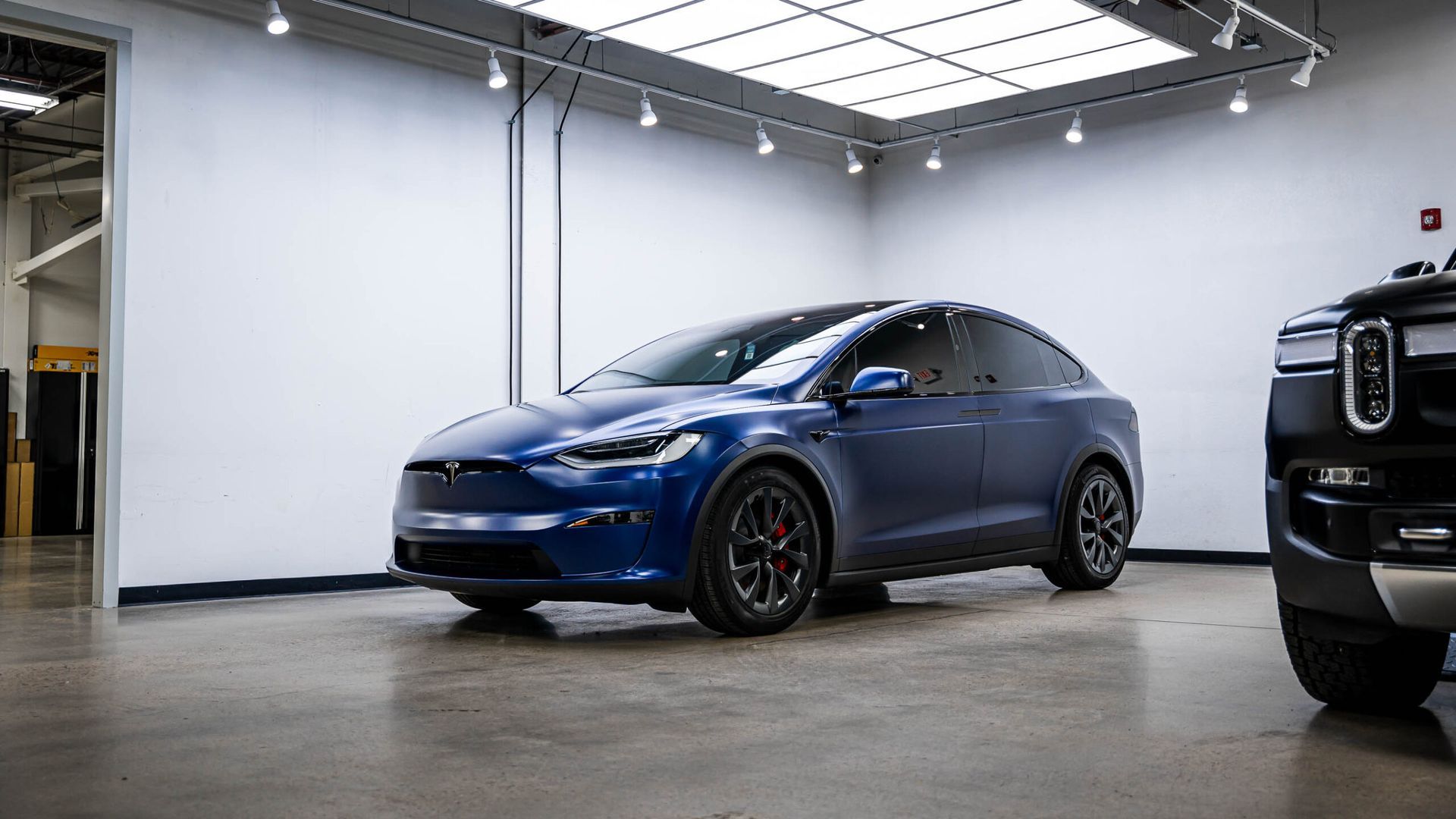 A blue tesla model x is parked in a garage next to a black truck.