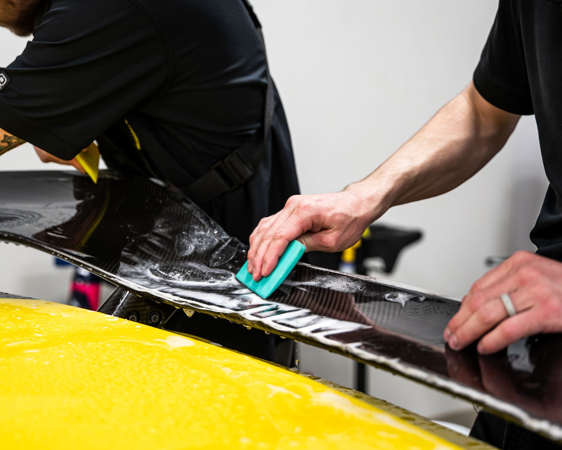Two people applying a film to a black spoiler on a yellow car using squeegees.
