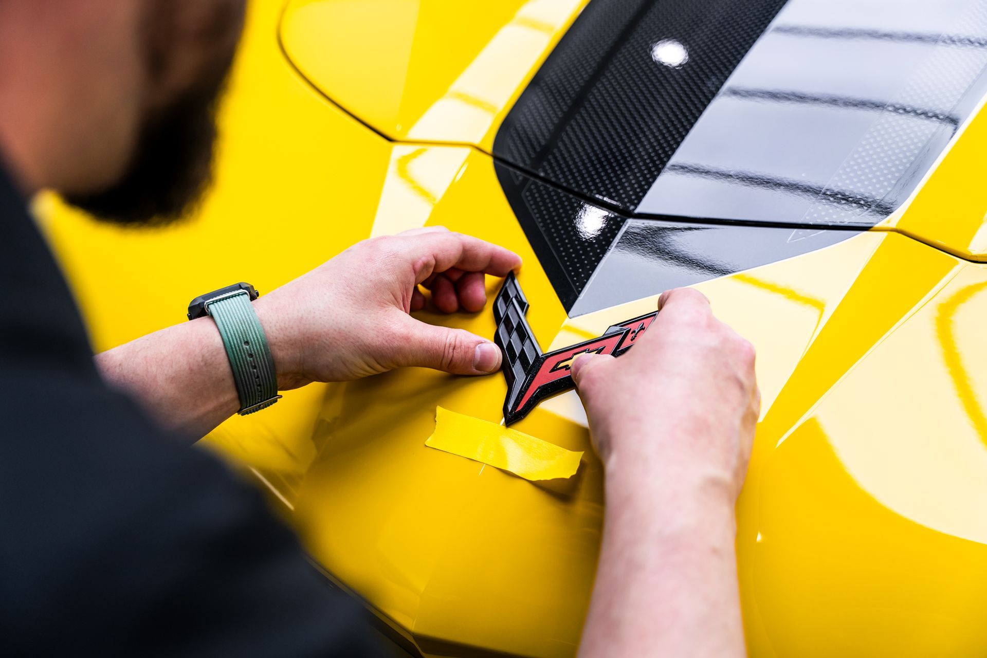 Person applying a red and black Corvette logo to a yellow car's hood.