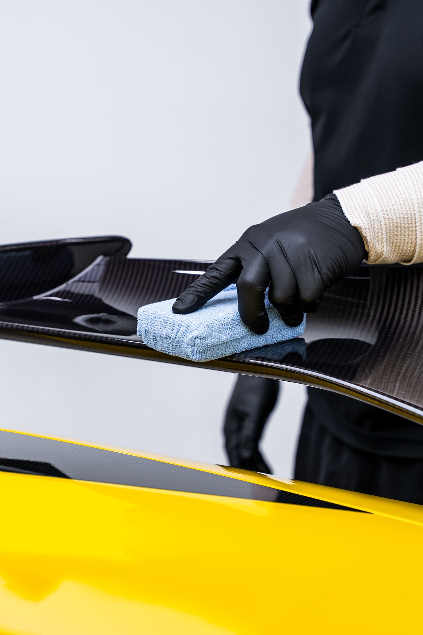A person wearing black gloves cleans a yellow sports car wing with a blue sponge.