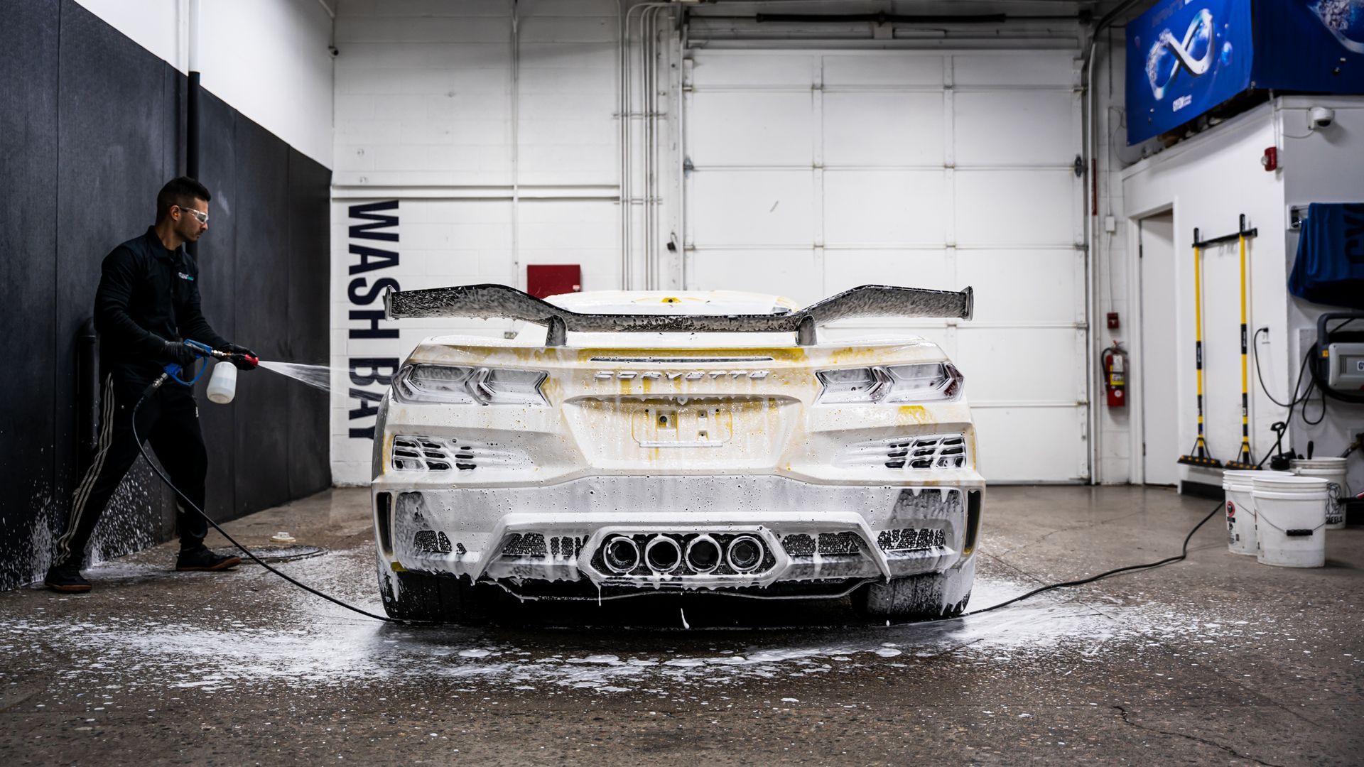 A person washes a white sports car in a wash bay, covered in foam.