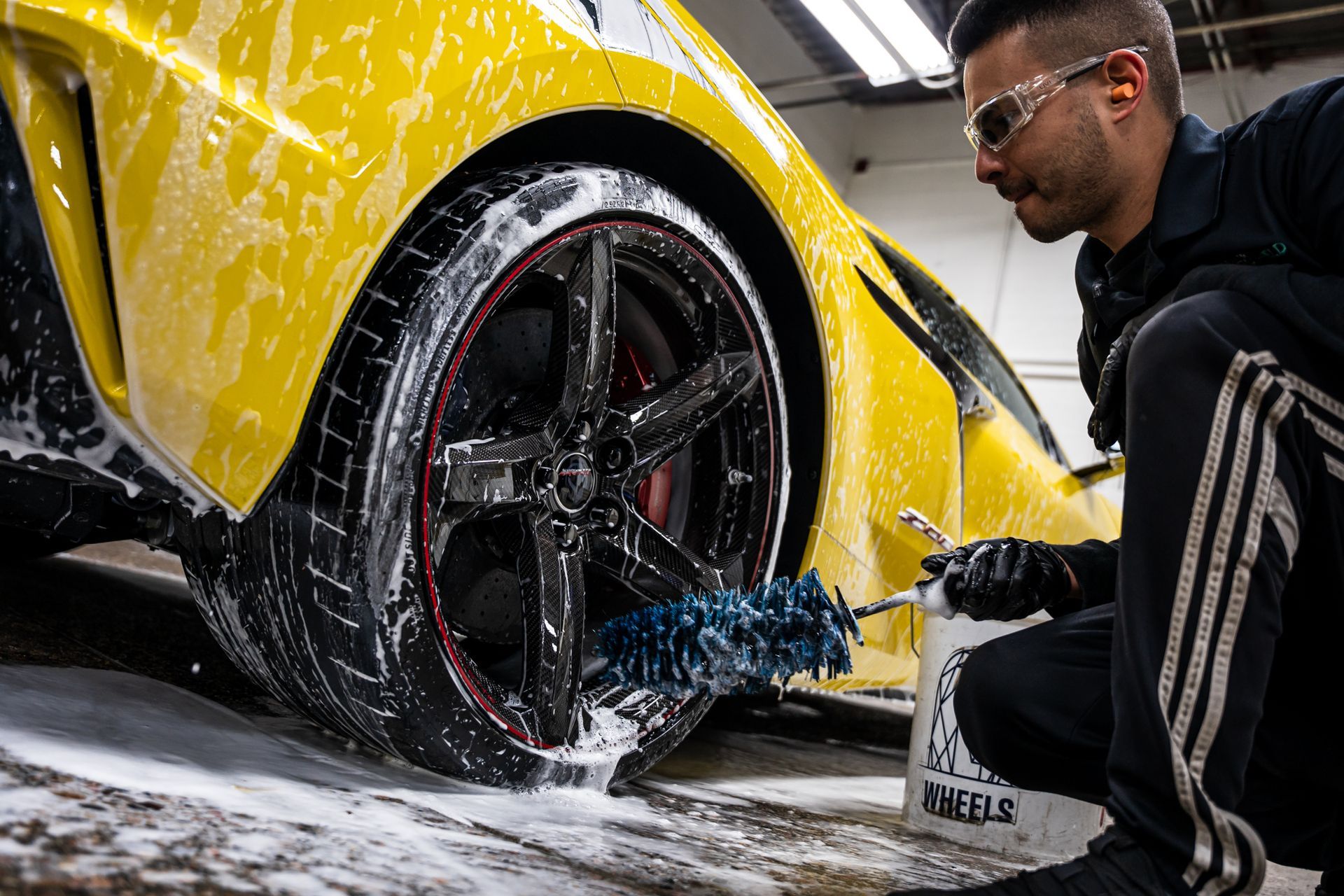 Man washing a yellow sports car's tire, wearing safety glasses, in a car wash bay.