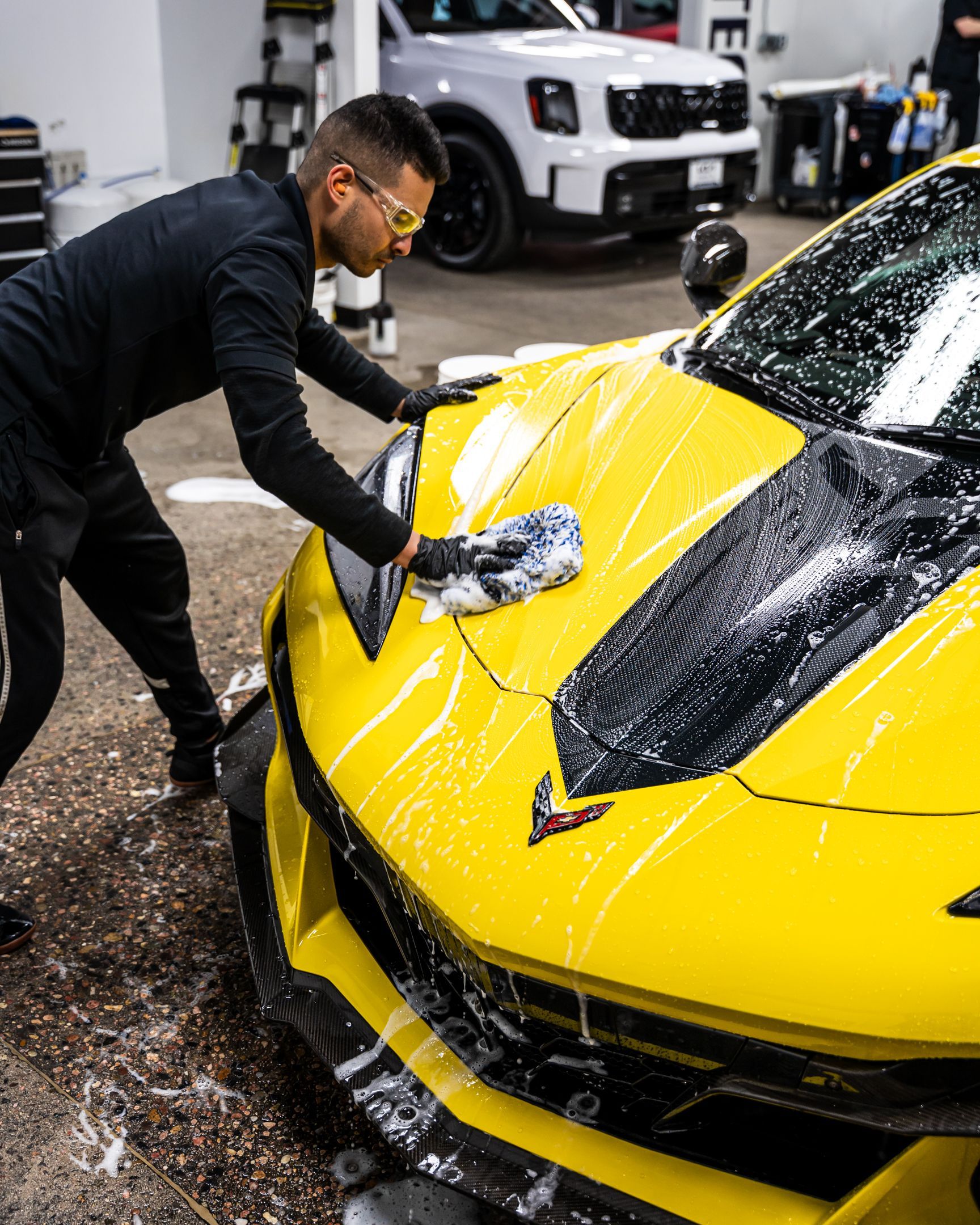 Man washes a bright yellow sports car, suds covering the hood, in a garage.