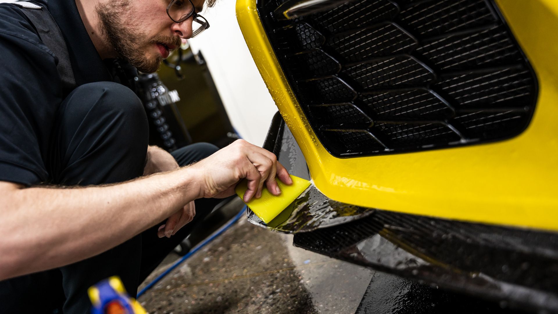Man applying film to a yellow car bumper with a squeegee in a garage setting.