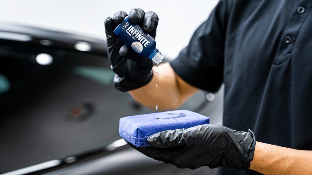A man wearing black gloves is holding a blue sponge and a bottle of ceramic coating.