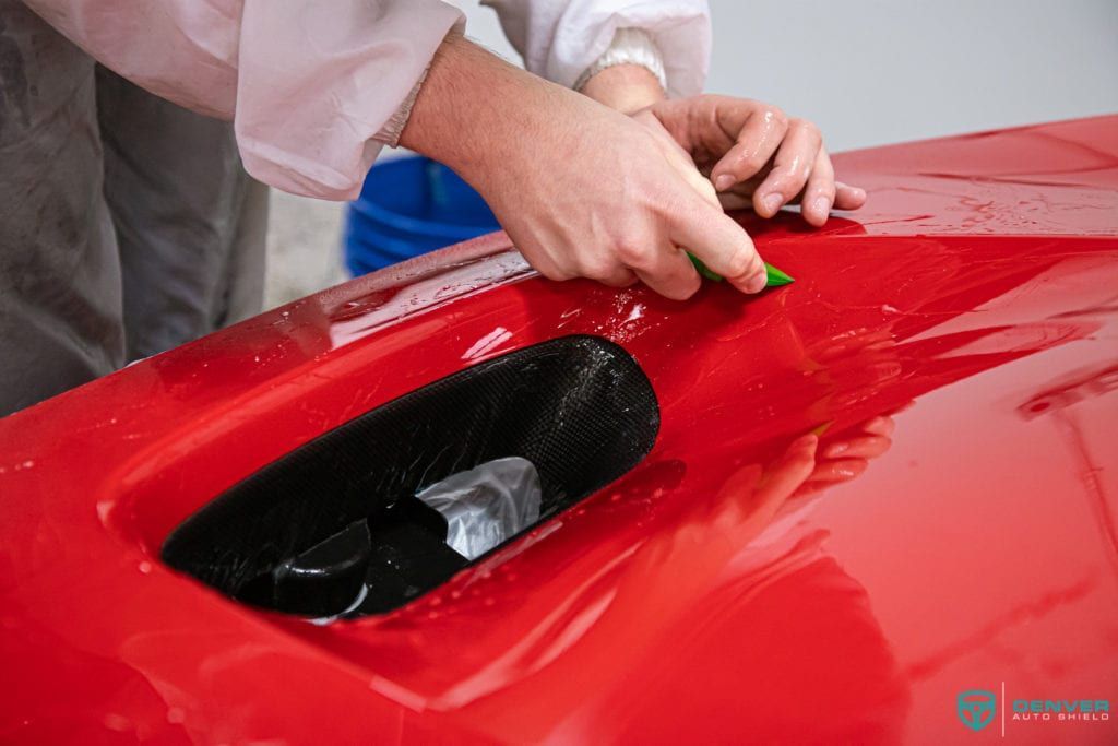 A person is applying a protective film to the hood of a red car.