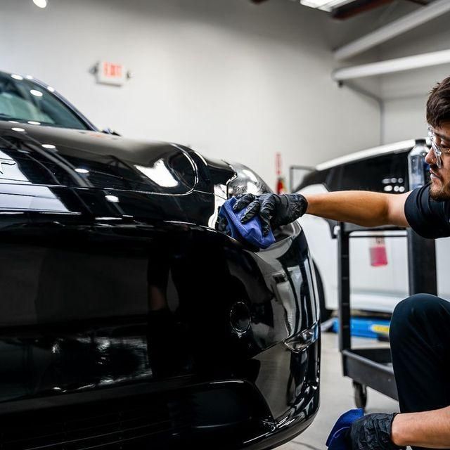 Detailing technician polishing a black car’s front bumper in a garage