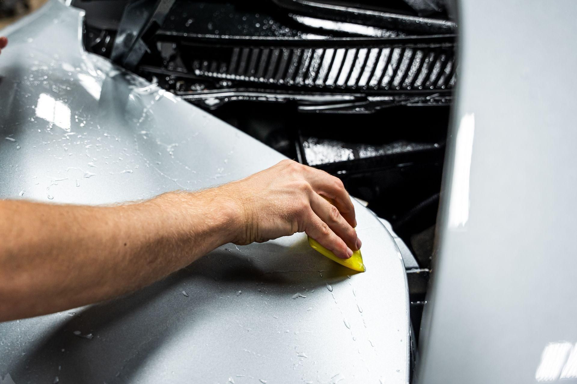 Hand polishing a white car hood with a yellow sponge near the windshield