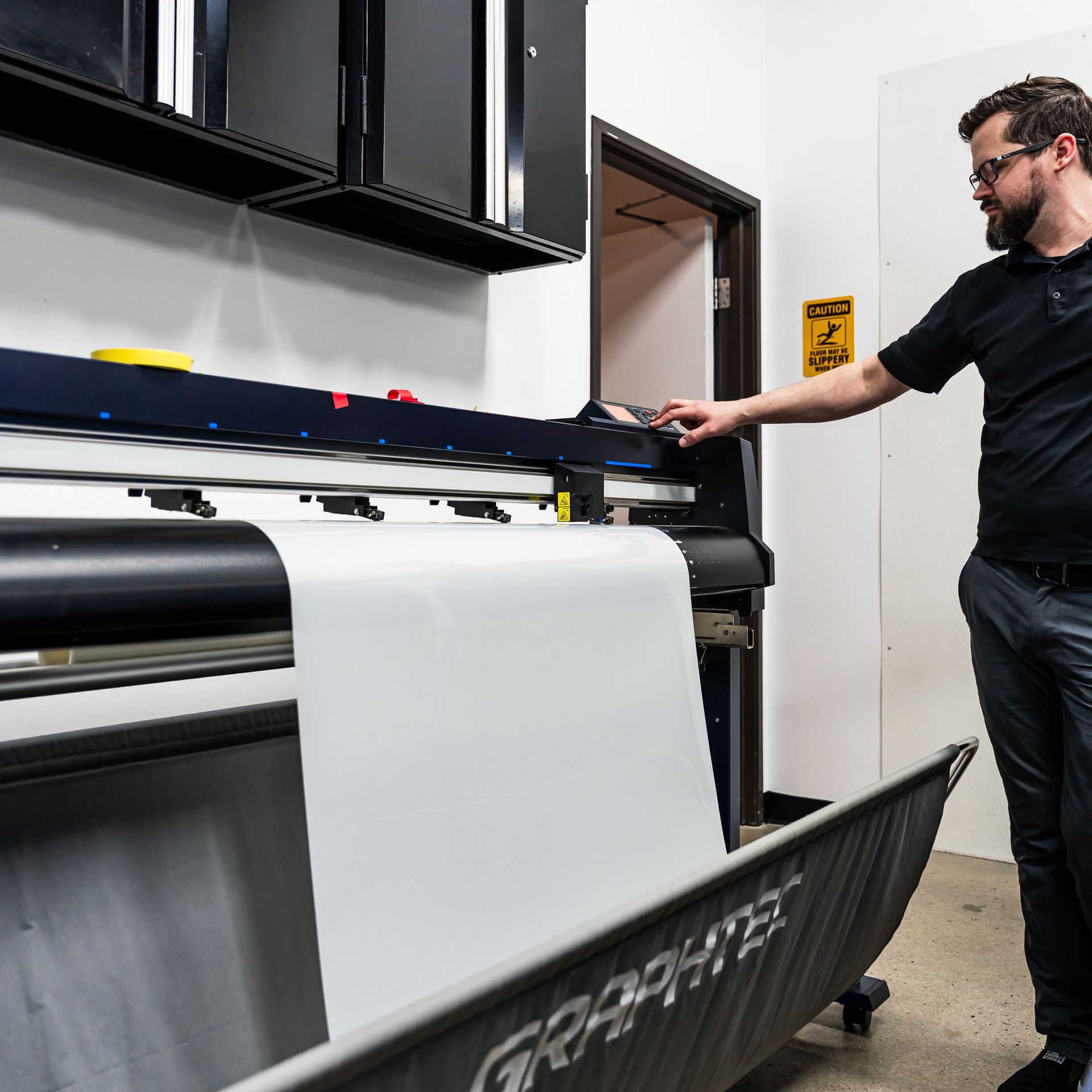 Man operating a large format printer in a print shop; white material rolls out.