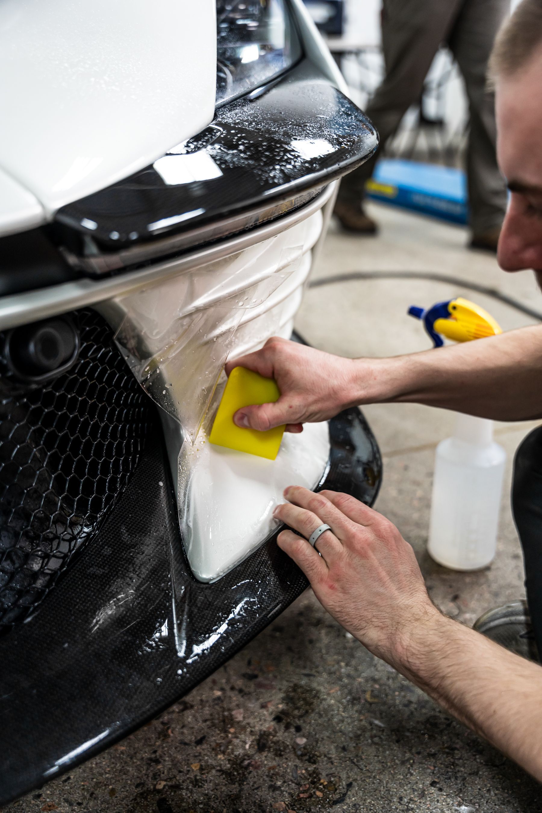 A man applying protective film to a car bumper using a squeegee in a workshop.