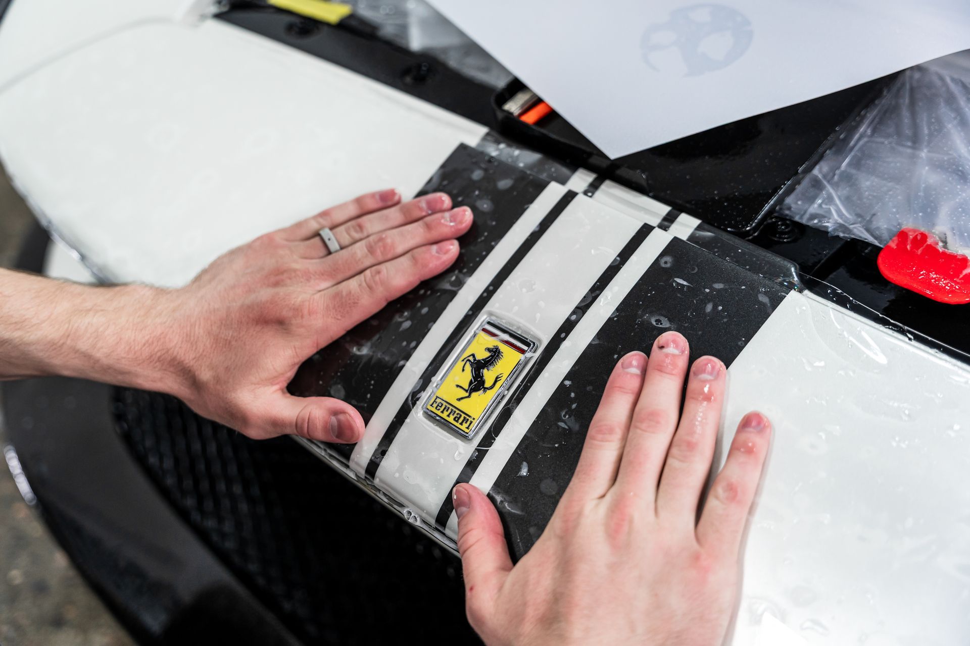 Hands applying a black and white racing stripe decal with a Ferrari logo on a white car hood.